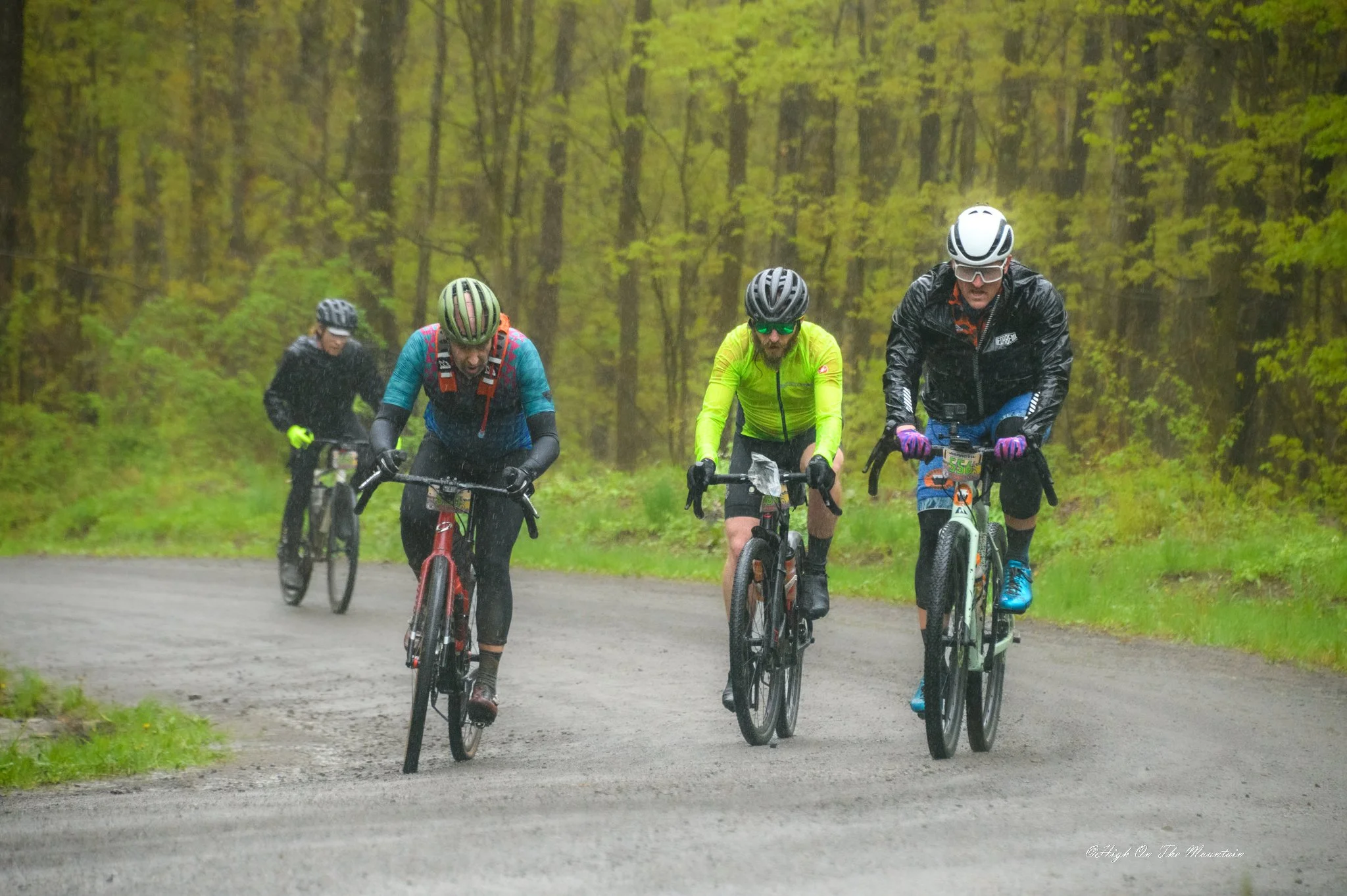 Four cyclists riding mountain bikes on a rainy forest trail, wearing helmets and rain gear.