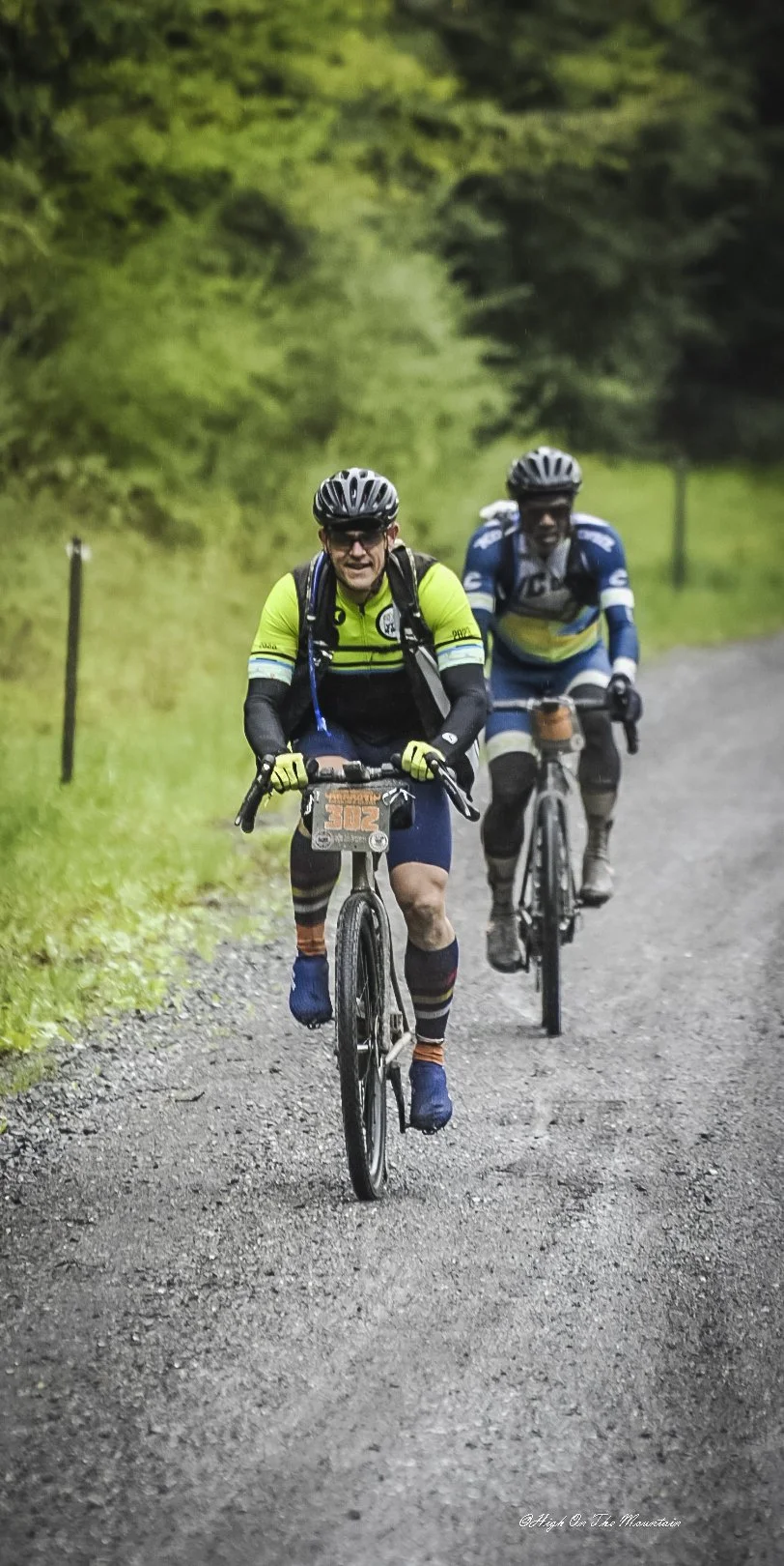 Two cyclists riding on a gravel trail through a wooded area, wearing helmets and cycling gear.