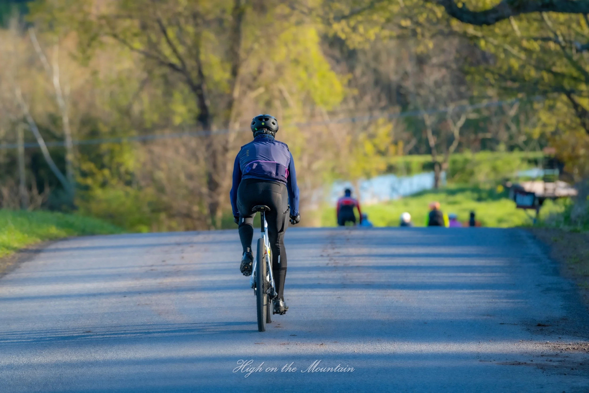 A person wearing a helmet biking on a paved road surrounded by green trees and grass, with other people sitting on the grass in the background.