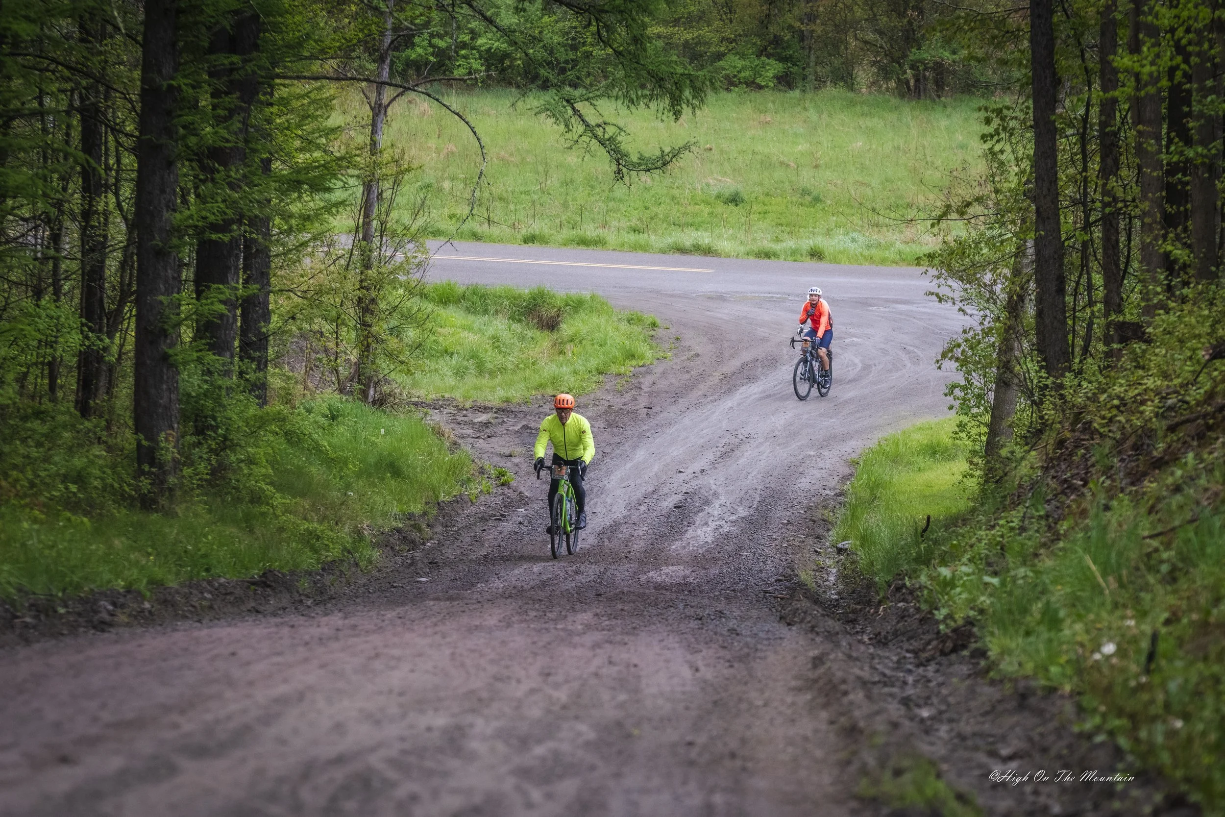 Two cyclists riding on a dirt trail through a lush green forest with a grassy area and a pavement road in the background.