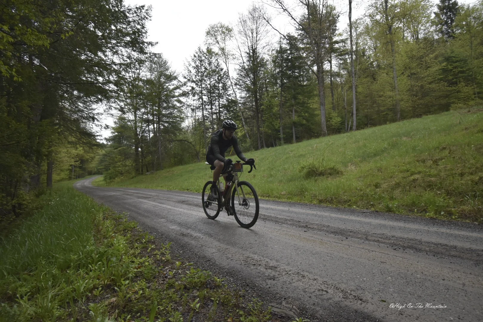 A cyclist riding a mountain bike on a gravel road through a forest with green trees and grass.