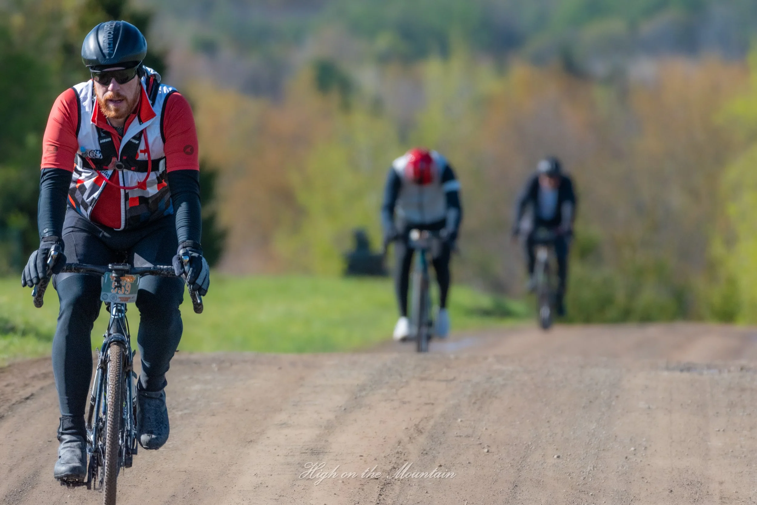A person wearing a helmet and sunglasses riding a mountain bike on a dirt trail, with two other cyclists in the background on a trail surrounded by trees and fall foliage.