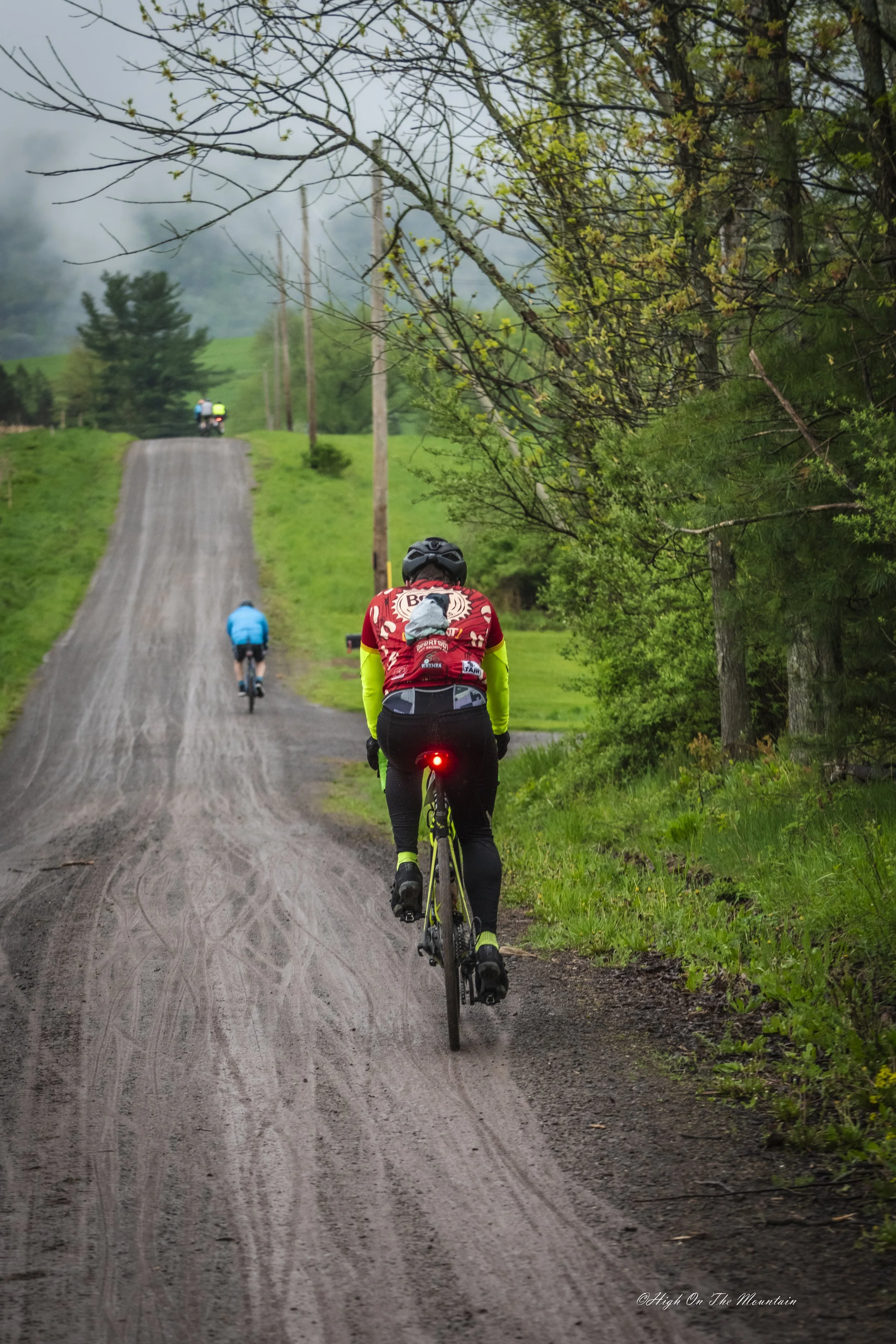 People riding bikes up a dirt trail in a lush, green, rural setting. The trail is surrounded by trees and grass, with some trees overhead and hills in the background.