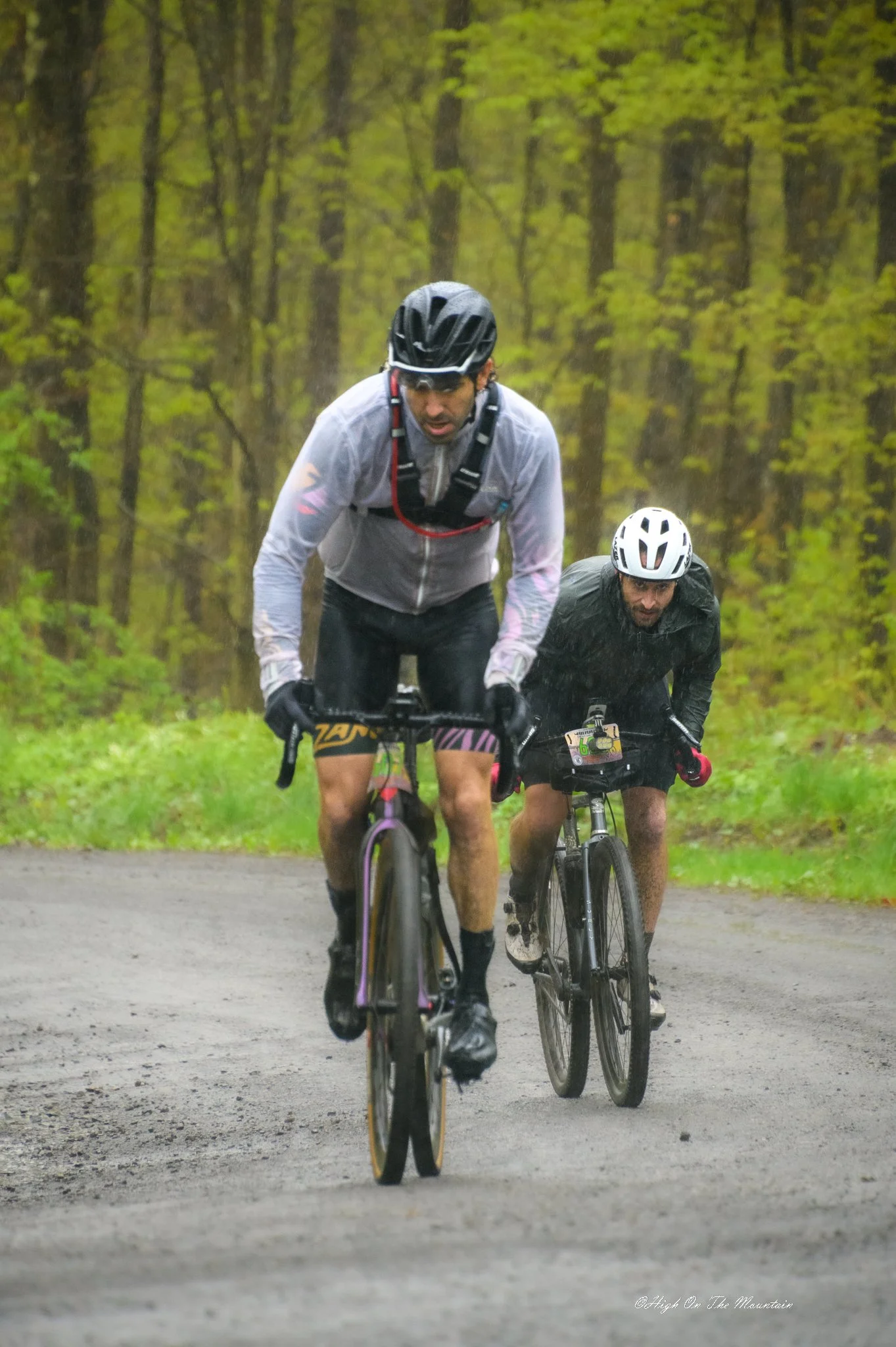 Two men riding bicycles on a dirt trail through a forest with green trees, wearing helmets and rain gear, with raindrops visible.