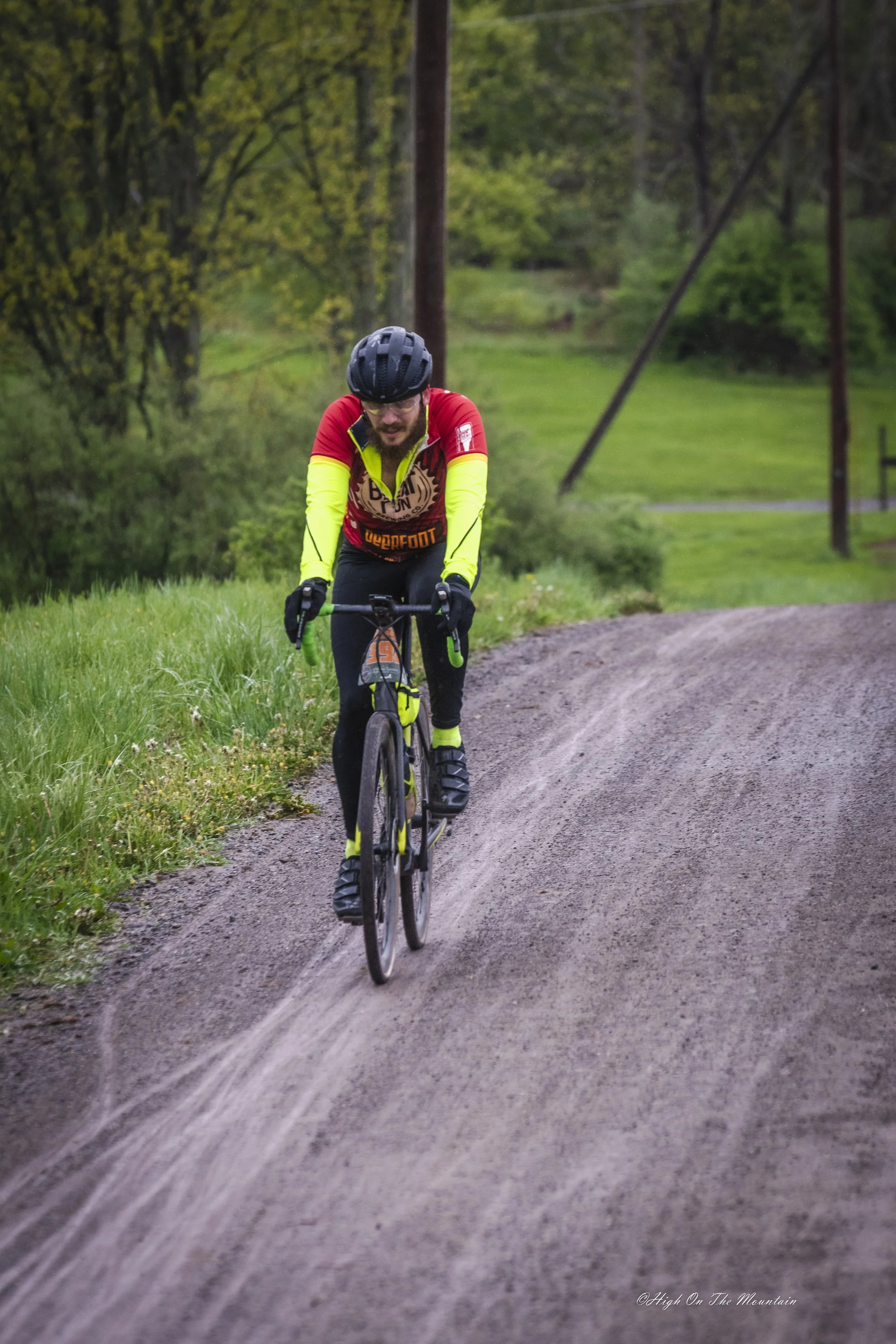 A person riding a mountain bike on a dirt trail through a green landscape with trees and utility poles in the background.