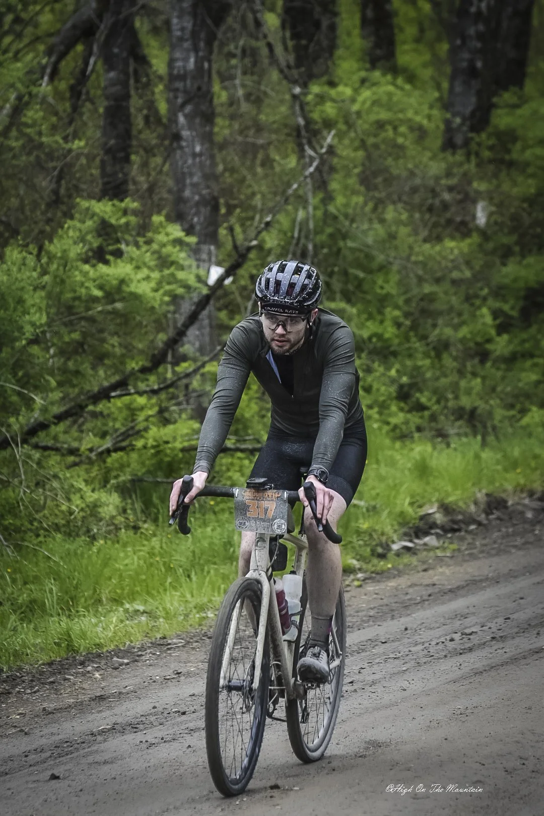 A male cyclist wearing a black helmet and glasses riding a mountain bike on a dirt trail through a green forest.