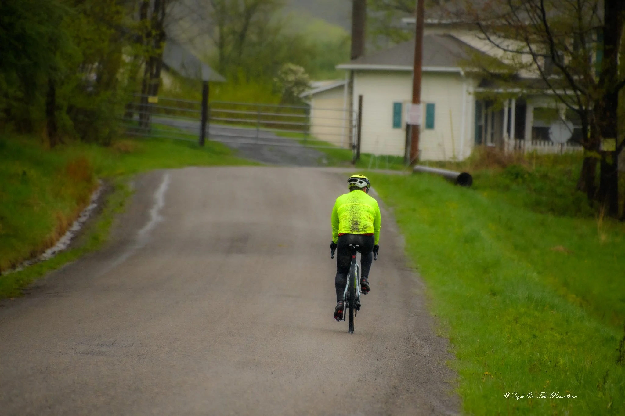 A person wearing a bright yellow jacket and helmet riding a bicycle on a paved country road lined with green grass and trees, with houses in the background.