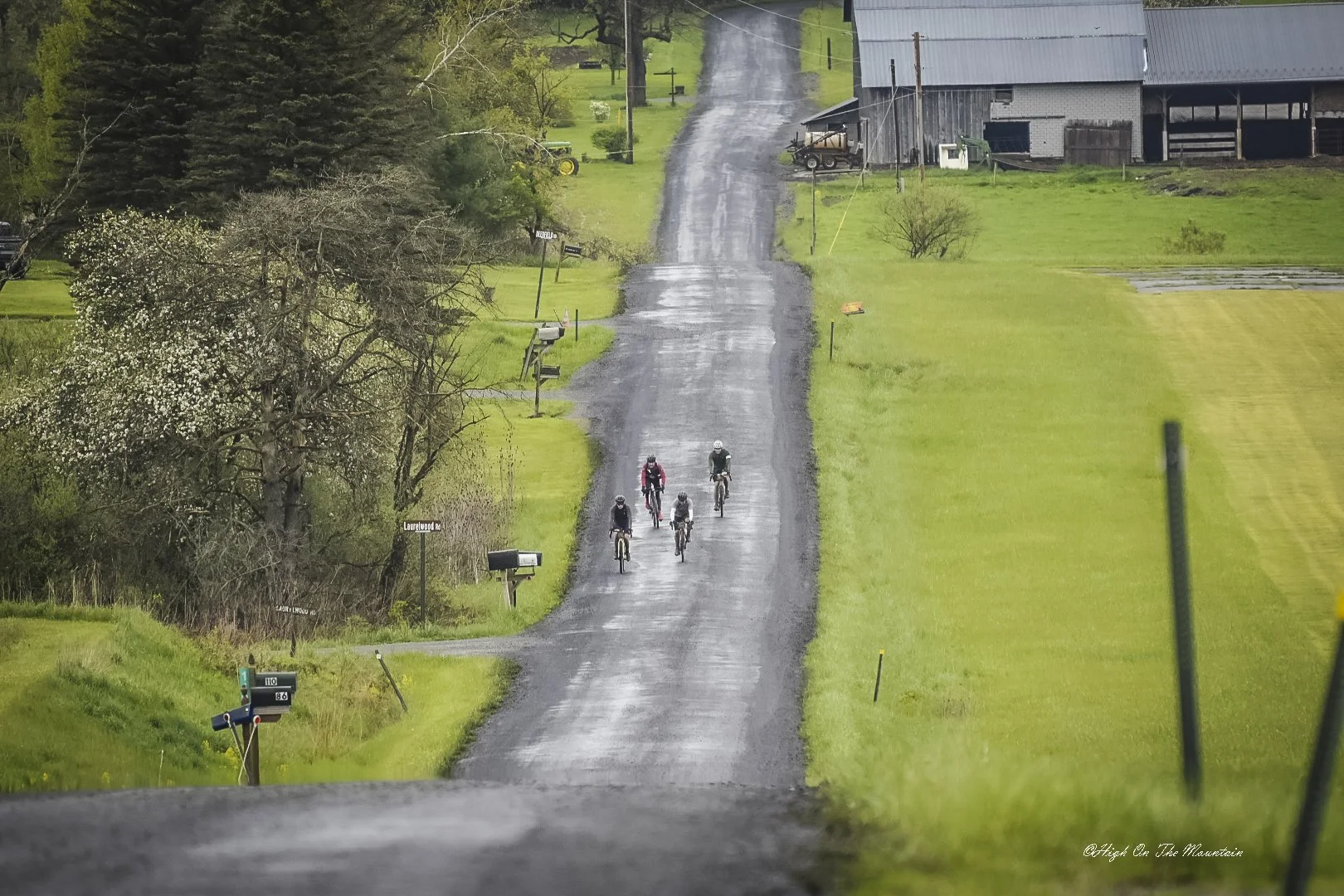 Four cyclists riding on a wet, rural gravel road surrounded by green grass and trees, with farm buildings in the background.