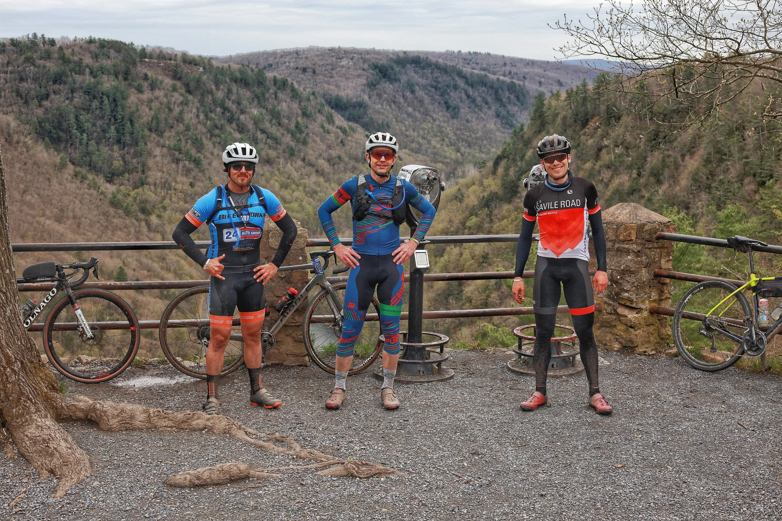 Three male cyclists in colorful cycling gear and helmets standing with their bikes on a mountain overlook with railing, overlooking a valley with hills and trees.