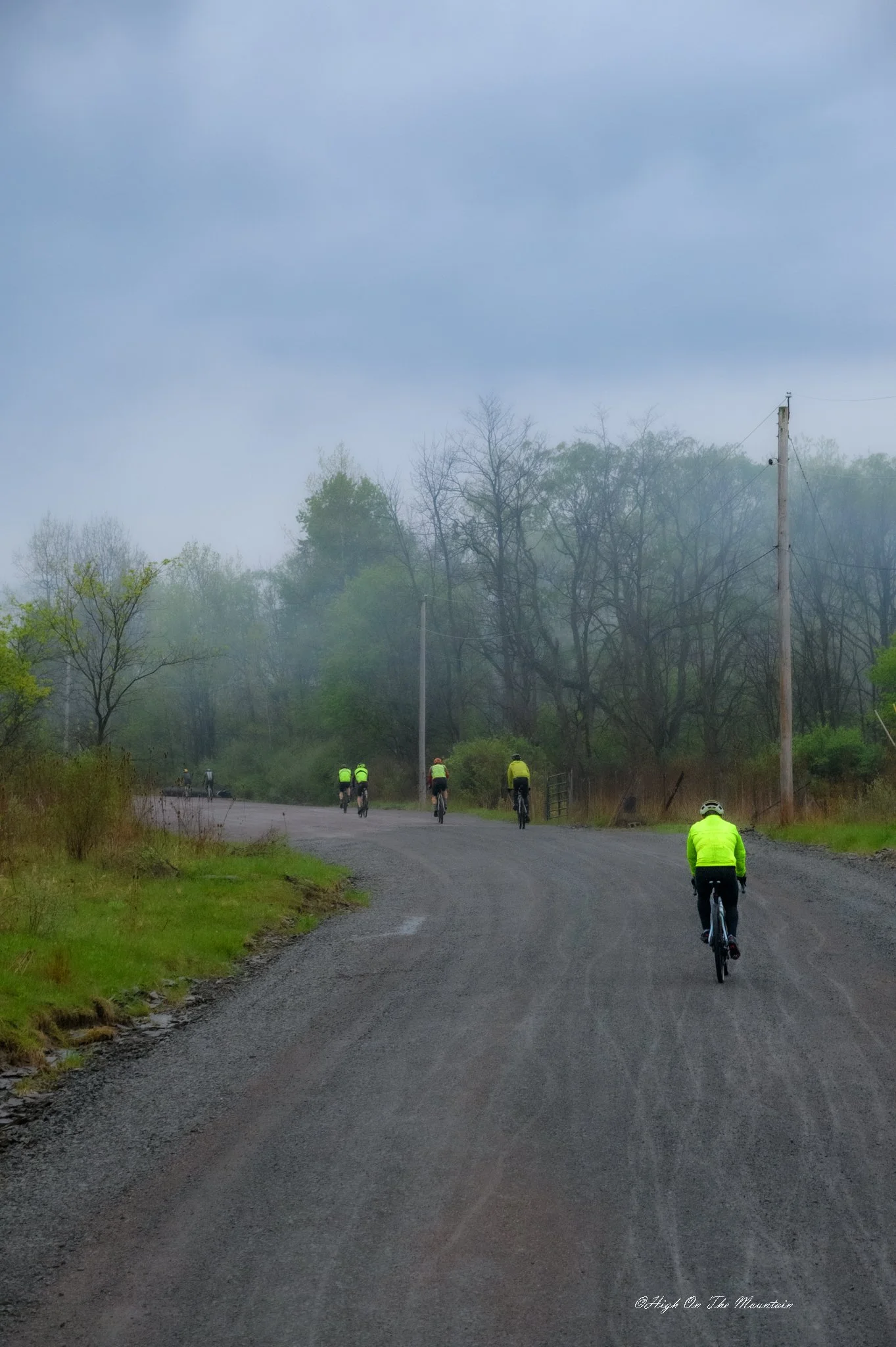 A group of cyclists dressed in bright yellow and green jackets riding on a gravel road surrounded by trees and overcast sky.
