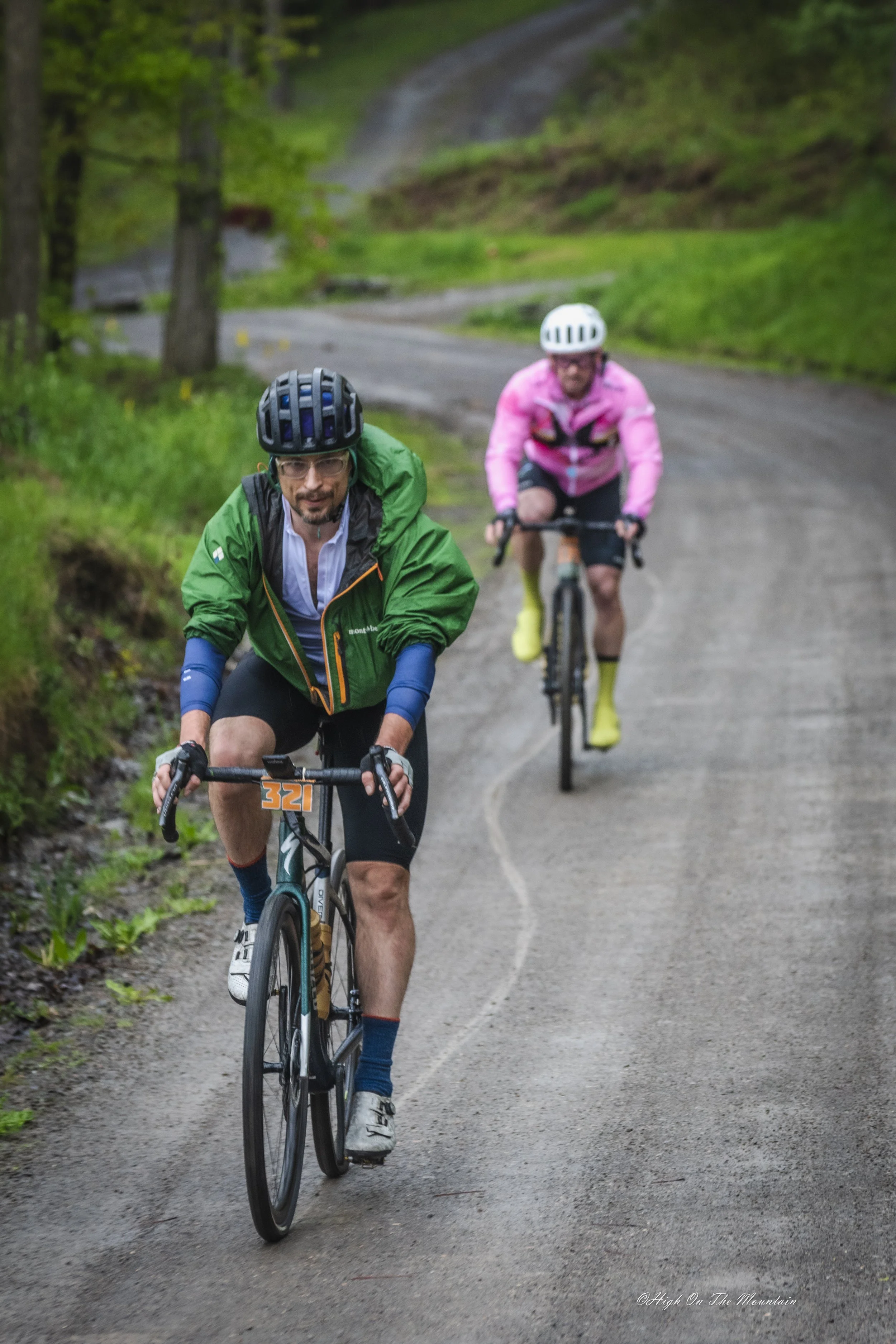 Two men riding mountain bikes on a winding forest trail. The man in the foreground wears a green jacket and black helmet, and the man behind him wears a pink jacket and white helmet. Both are equipped with cycling gear.