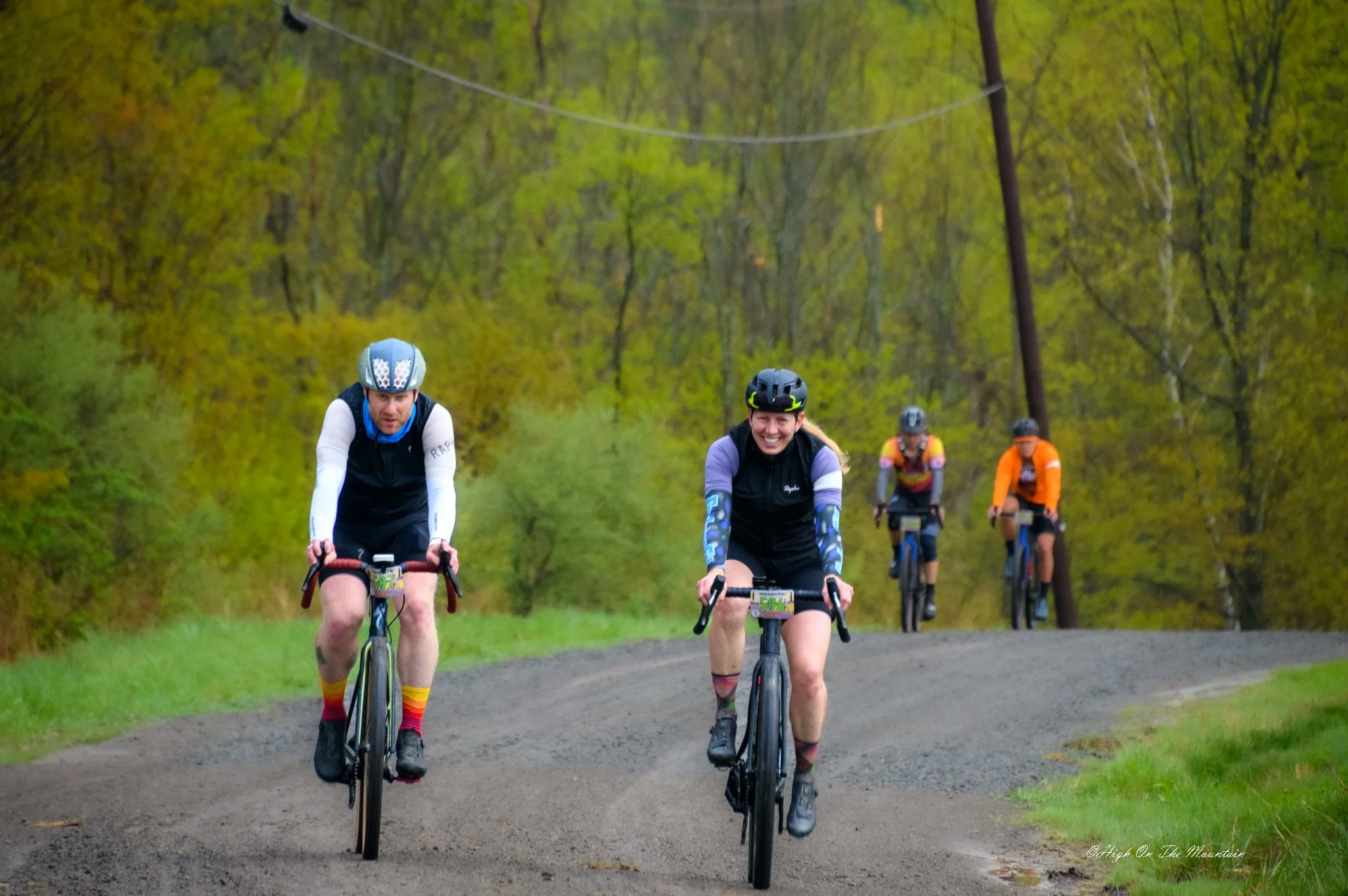 Four cyclists riding on a dirt trail through a green forest, some smiling and wearing helmets and colorful clothing.