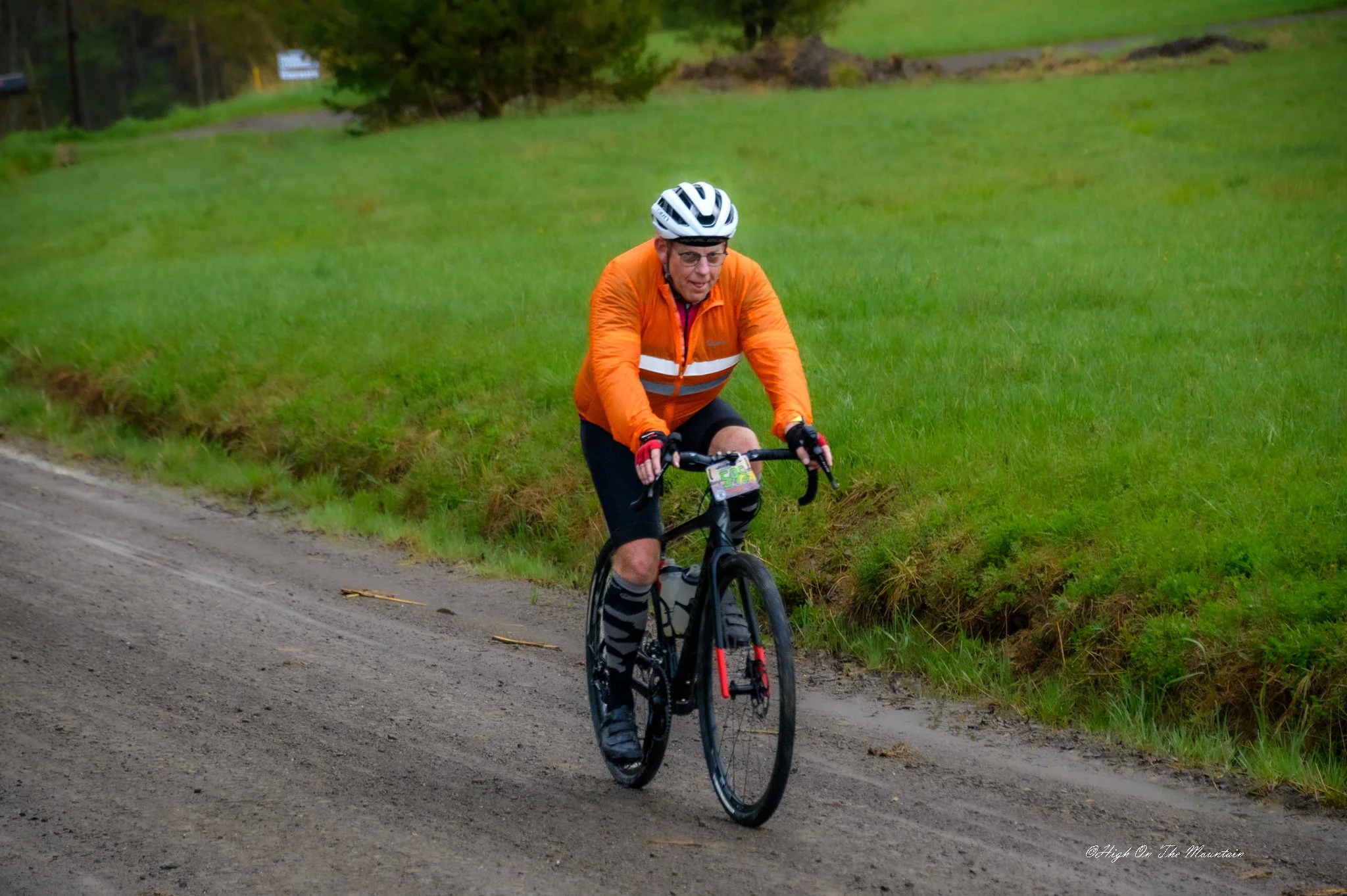 A man riding a mountain bike on a dirt trail through a grassy area on a rainy day, wearing an orange jacket, a white helmet, and glasses.