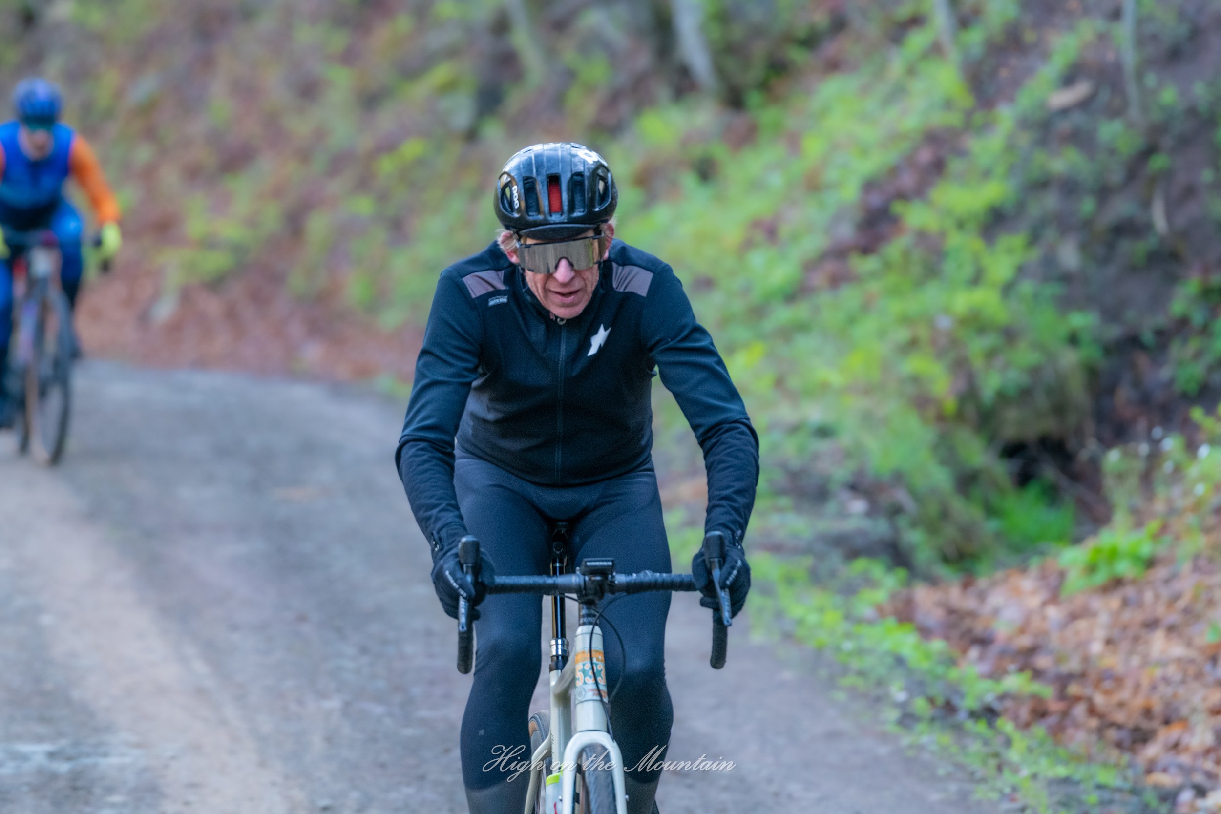 Two cyclists riding on a forest trail, with one cyclist in the foreground wearing a black helmet and sunglasses, and another cyclist in the background wearing an orange and blue outfit.