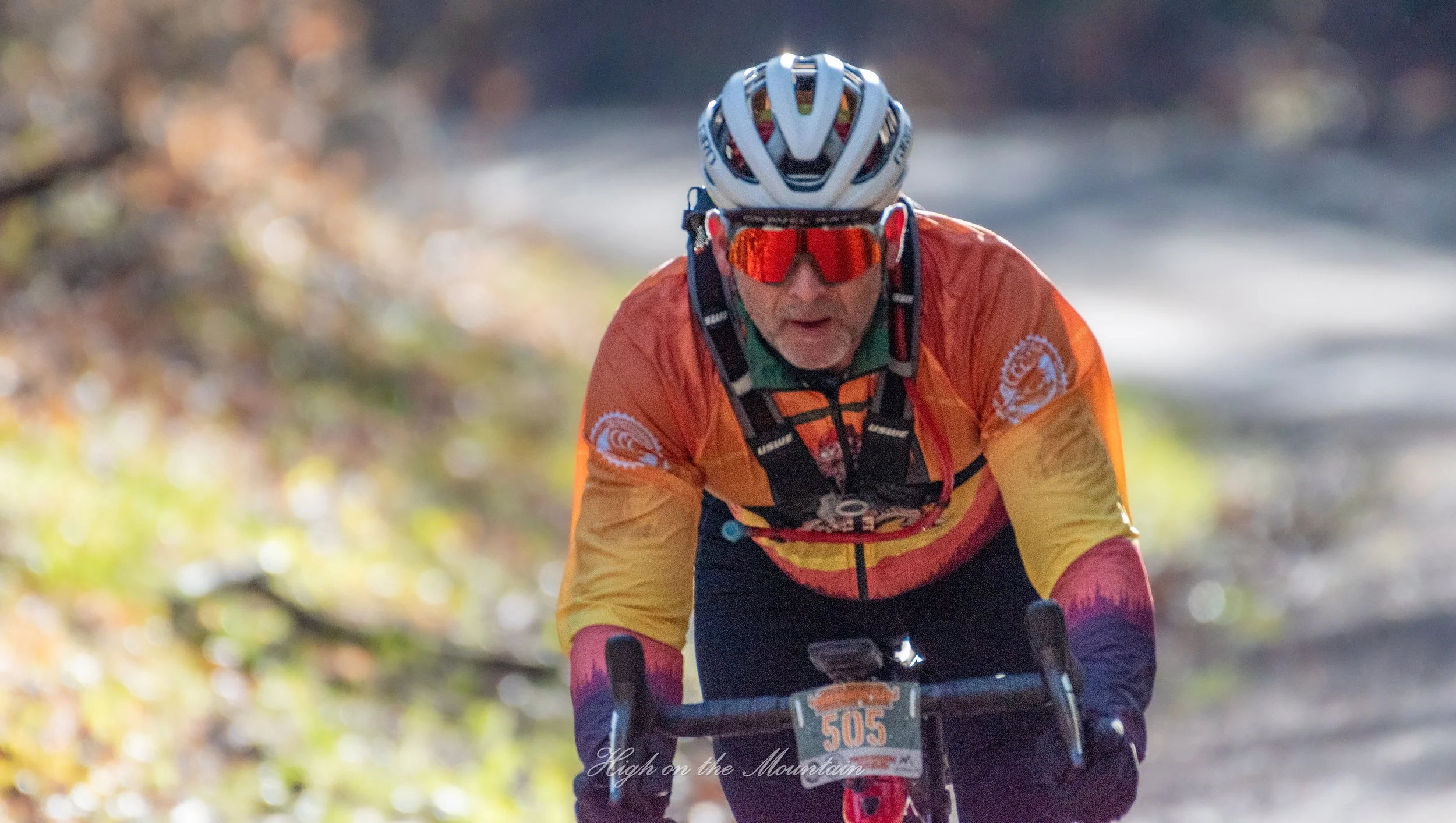 A cyclist wearing a white helmet, red-tinted goggles, and orange and pink cycling gear rides through a wooded area.