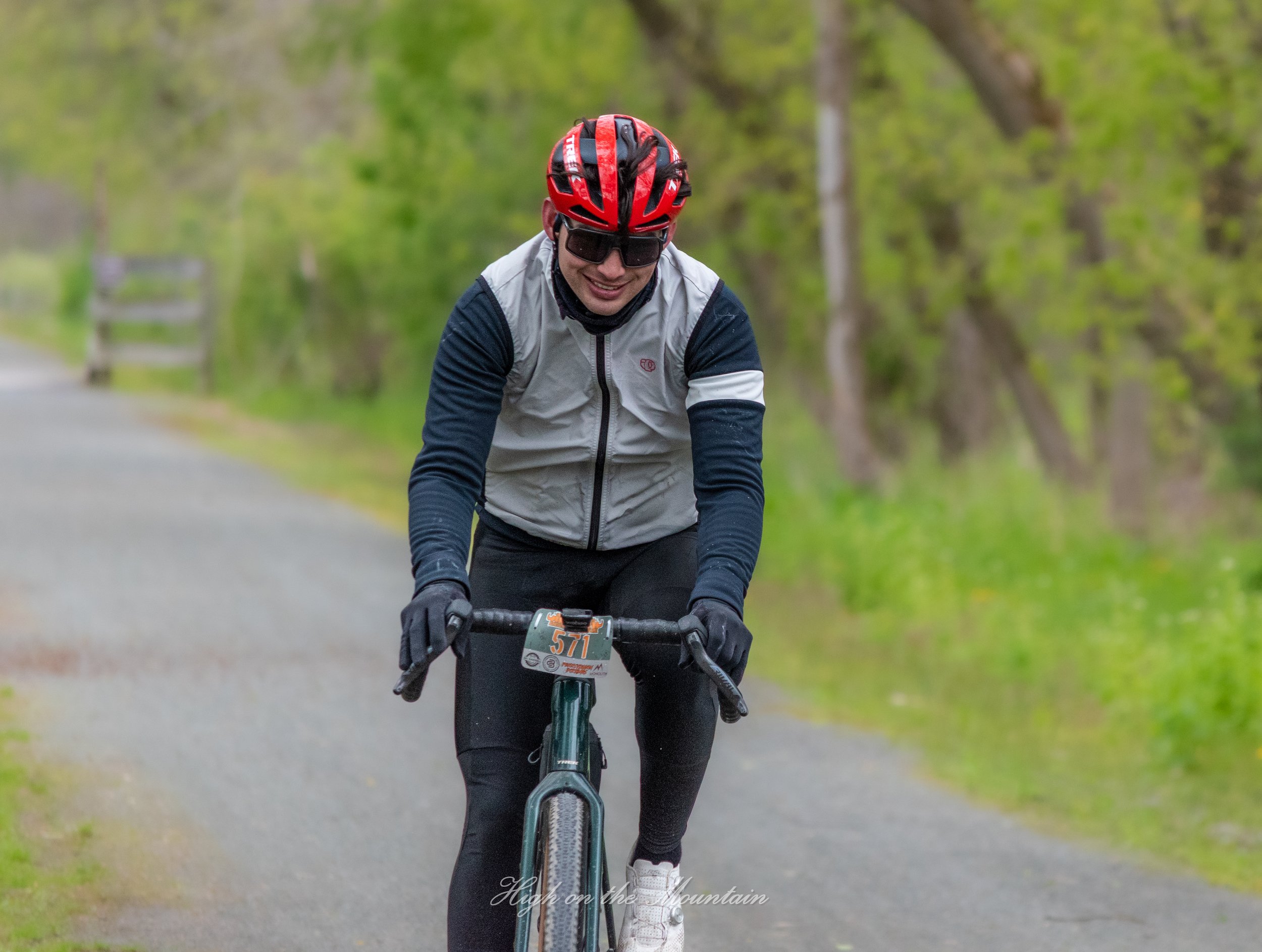 A person riding a mountain bike on a trail surrounded by green trees, wearing a red helmet, sunglasses, and a gray and black jacket.