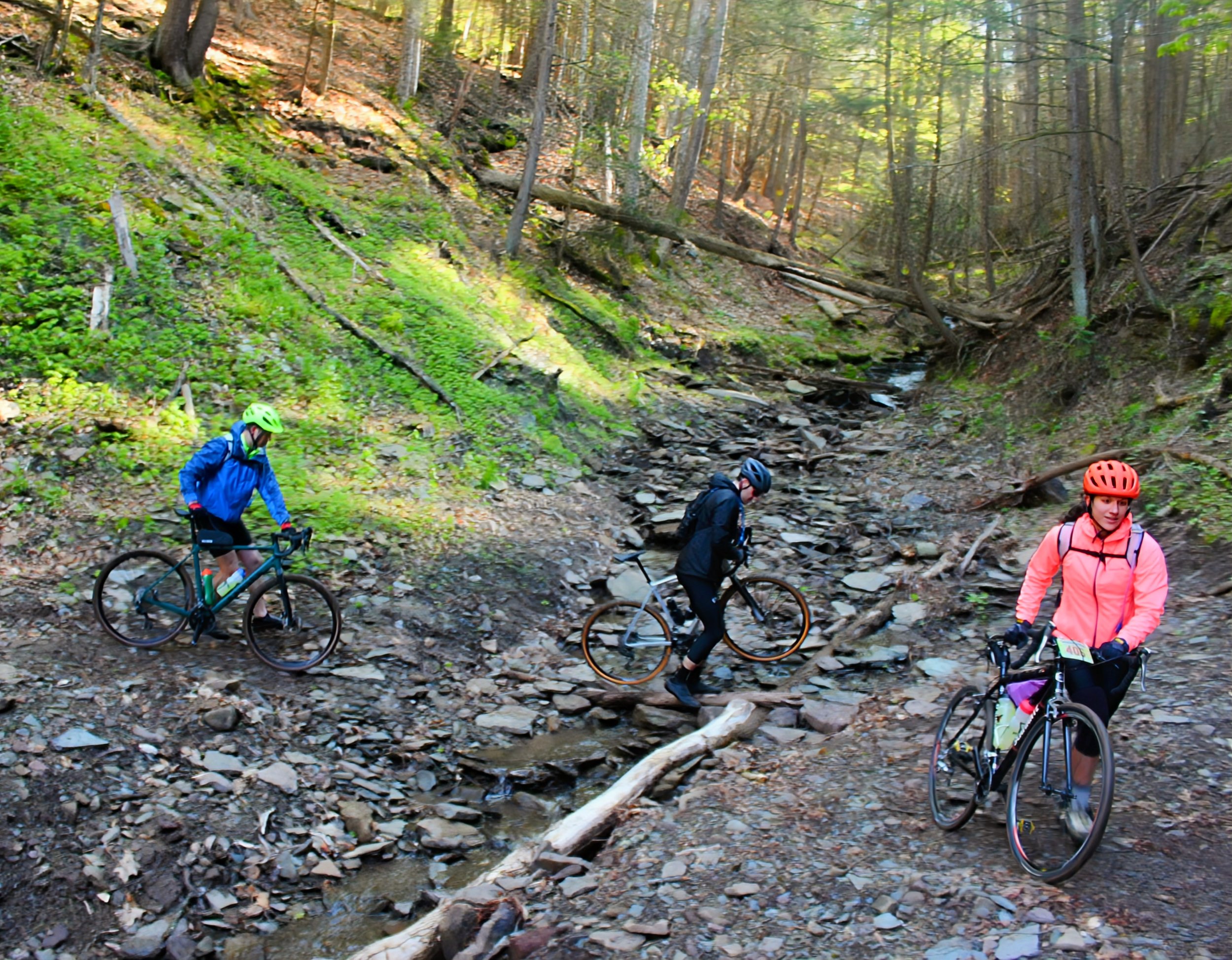 Three cyclists with helmets and gear crossing a rocky stream in a forest during daytime.