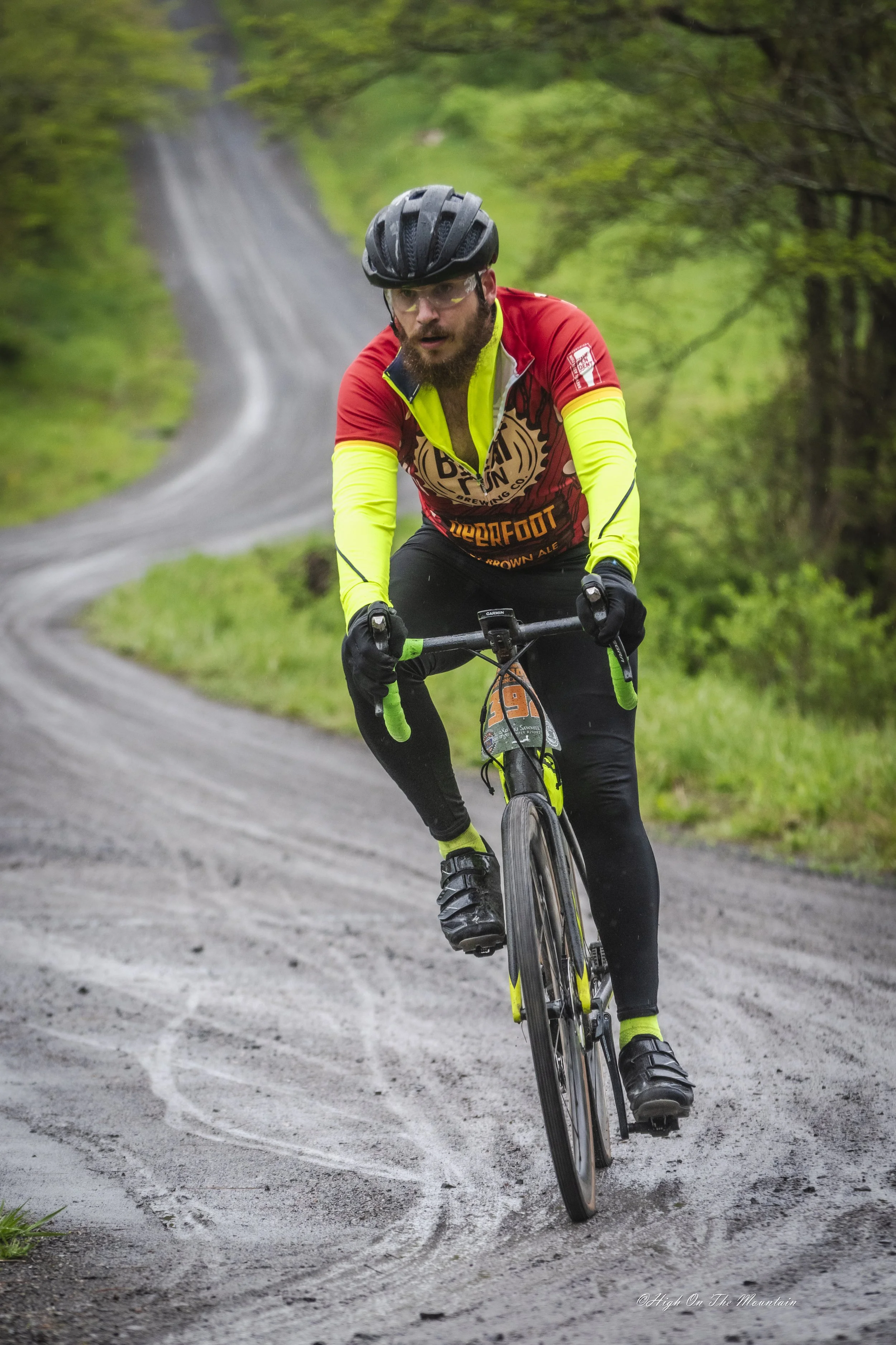 A man wearing a black helmet, yellow sunglasses, a bright yellow and red cycling jersey, black pants, and gloves riding a cycling bike on a winding dirt trail surrounded by green trees and foliage.
