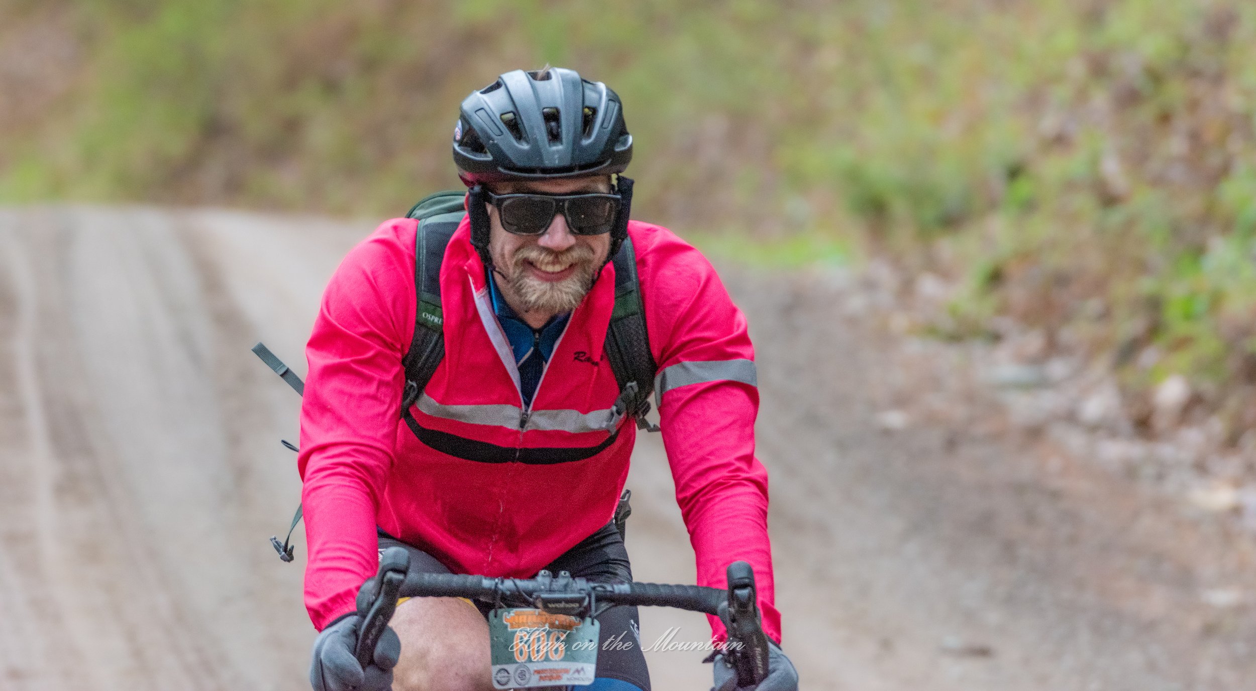 A man wearing a black helmet, dark sunglasses, and a bright pink jacket riding a mountain bike on a dirt trail surrounded by greenery.
