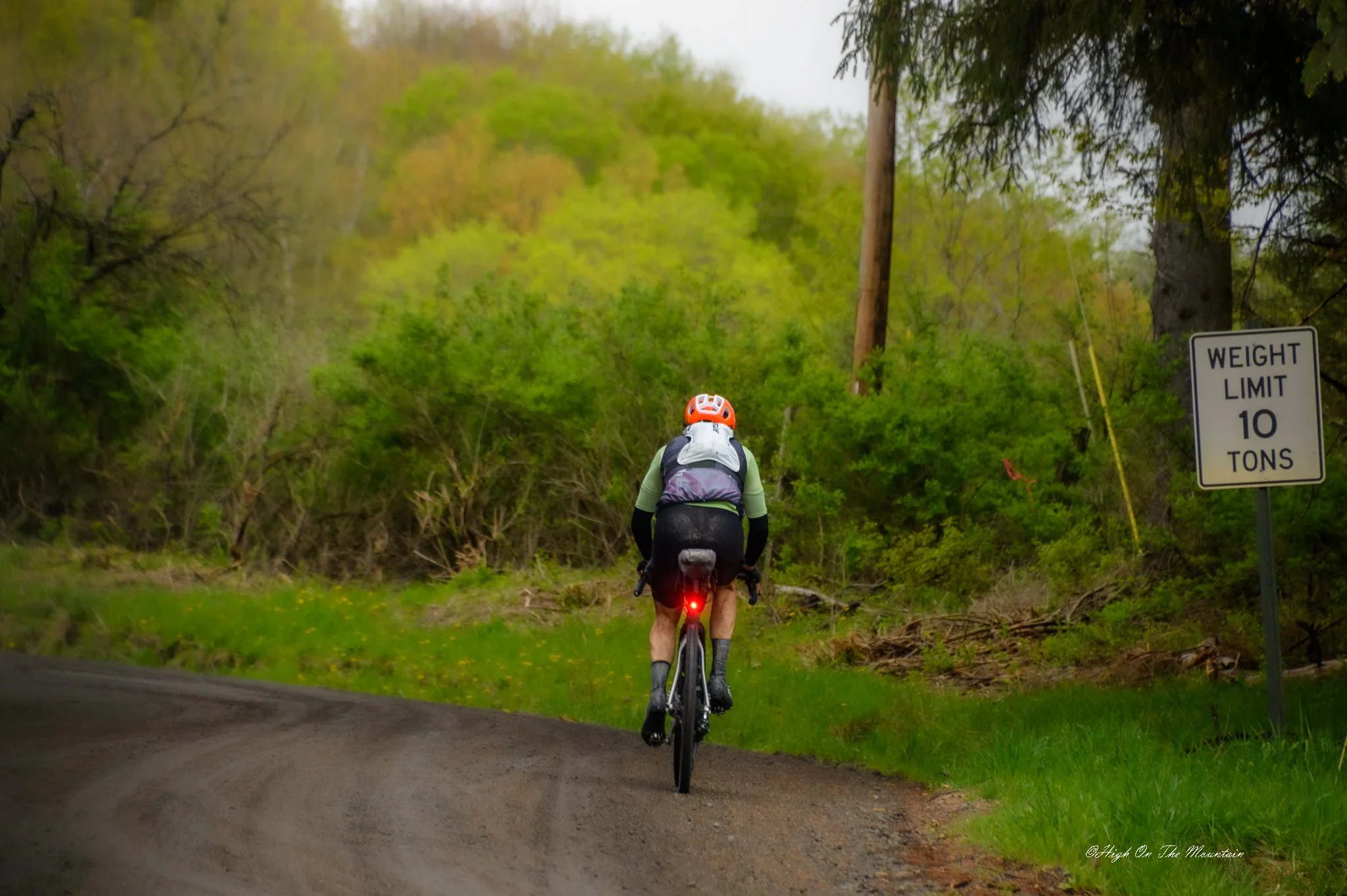 A person riding a mountain bike down a dirt path surrounded by green trees and foliage. The cyclist is wearing a white helmet, black shorts, and a light green long-sleeve shirt. A sign on the right indicates a weight limit of 10 tons.