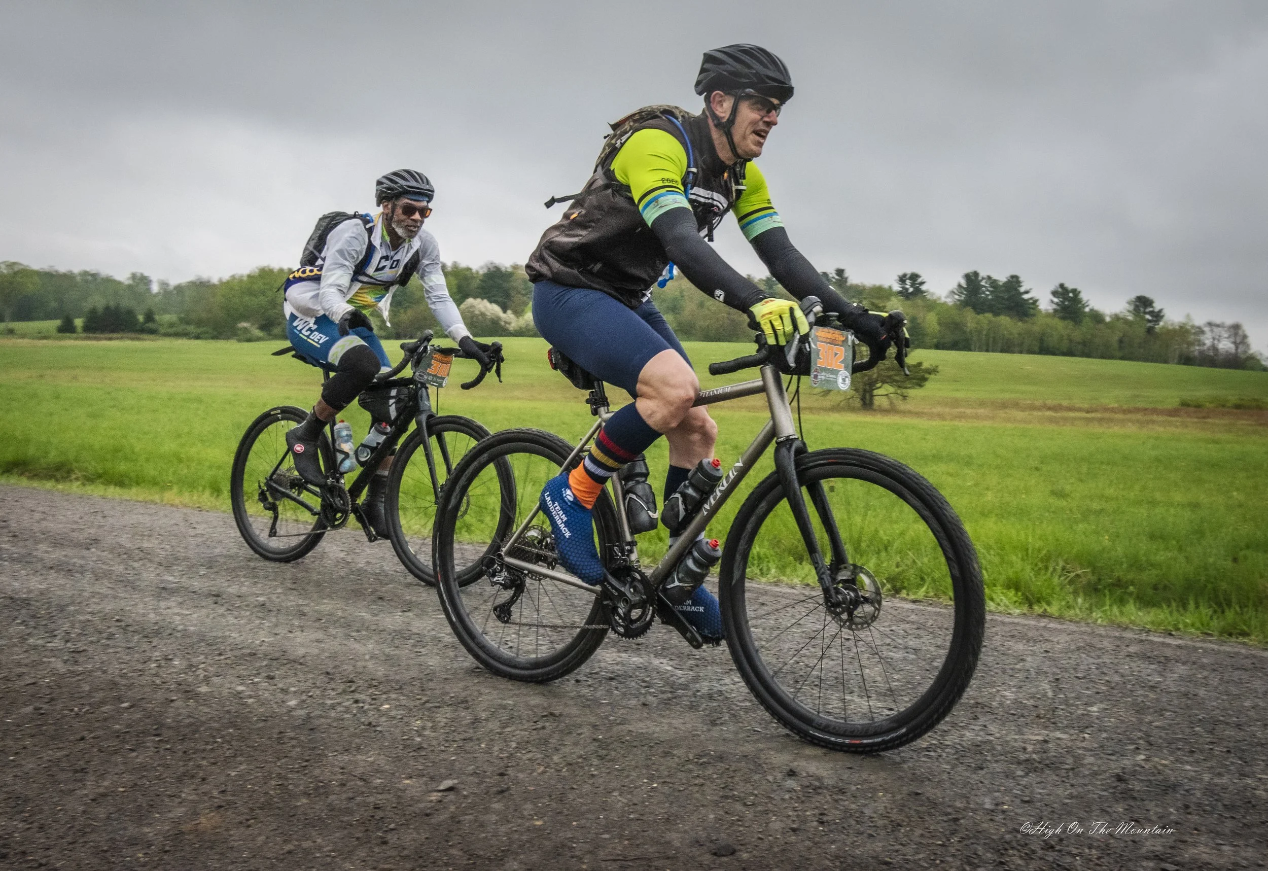 Two men riding bicycles on a gravel road in a grassy field with cloudy skies, wearing cycling gear and helmets.