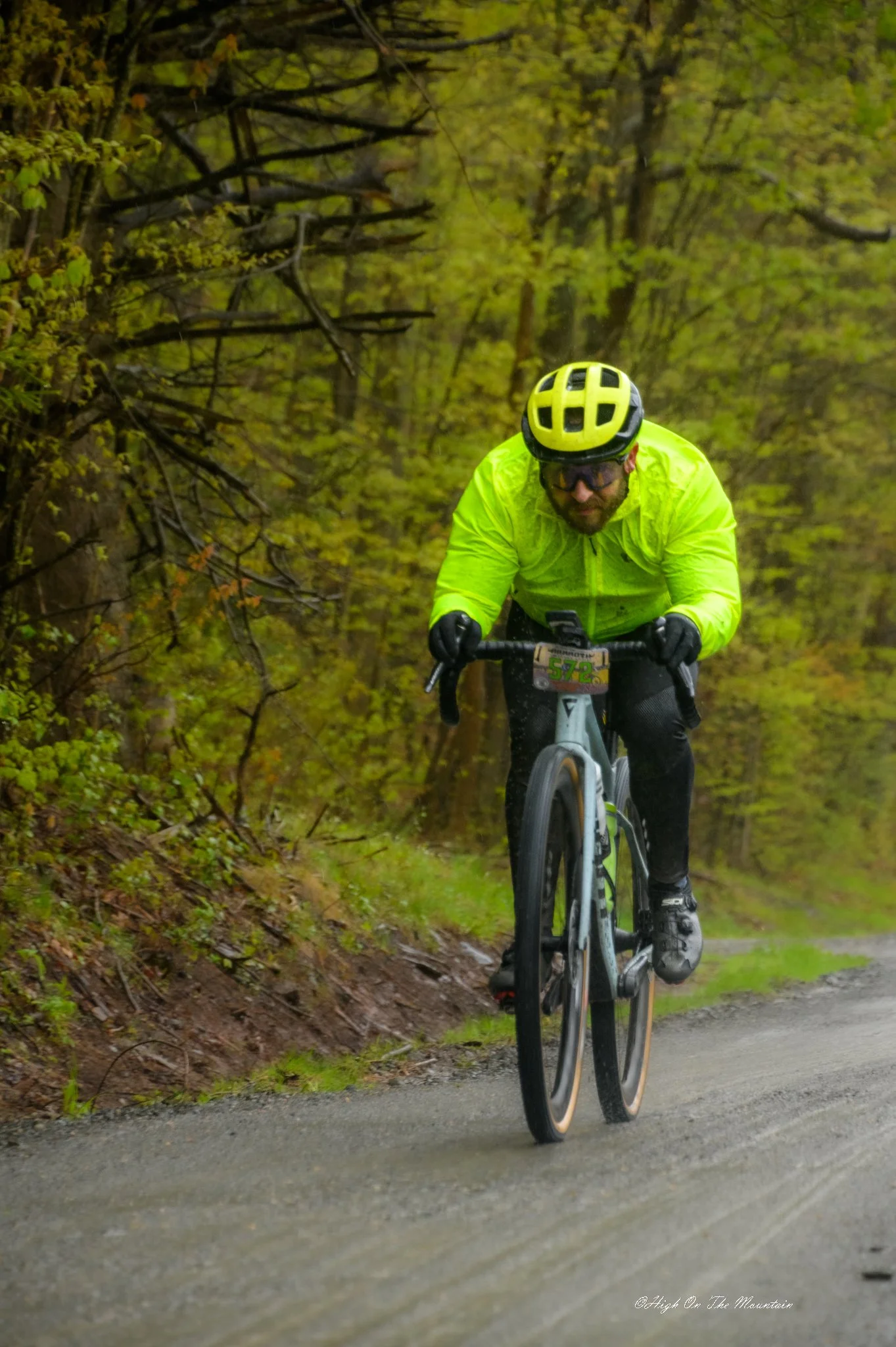 A mountain biker wearing a yellow jacket and helmet riding on a wet trail through a forest with green foliage.