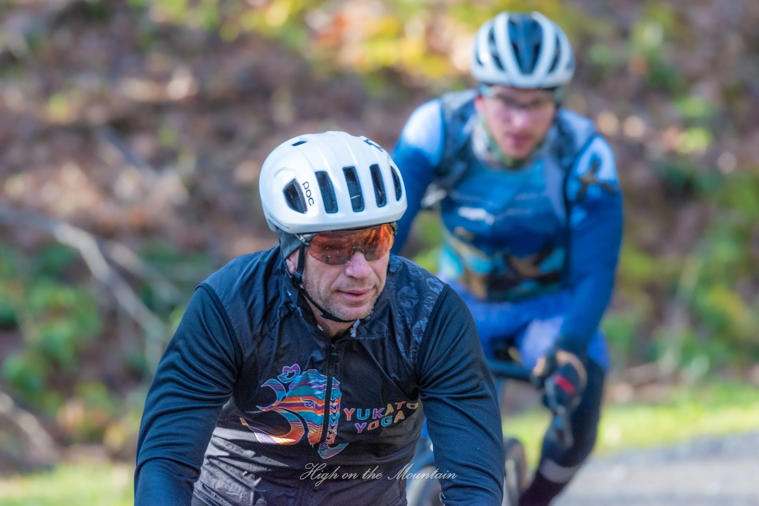 Two men wearing helmets and biking gear, riding mountain bikes on a trail surrounded by trees and foliage.