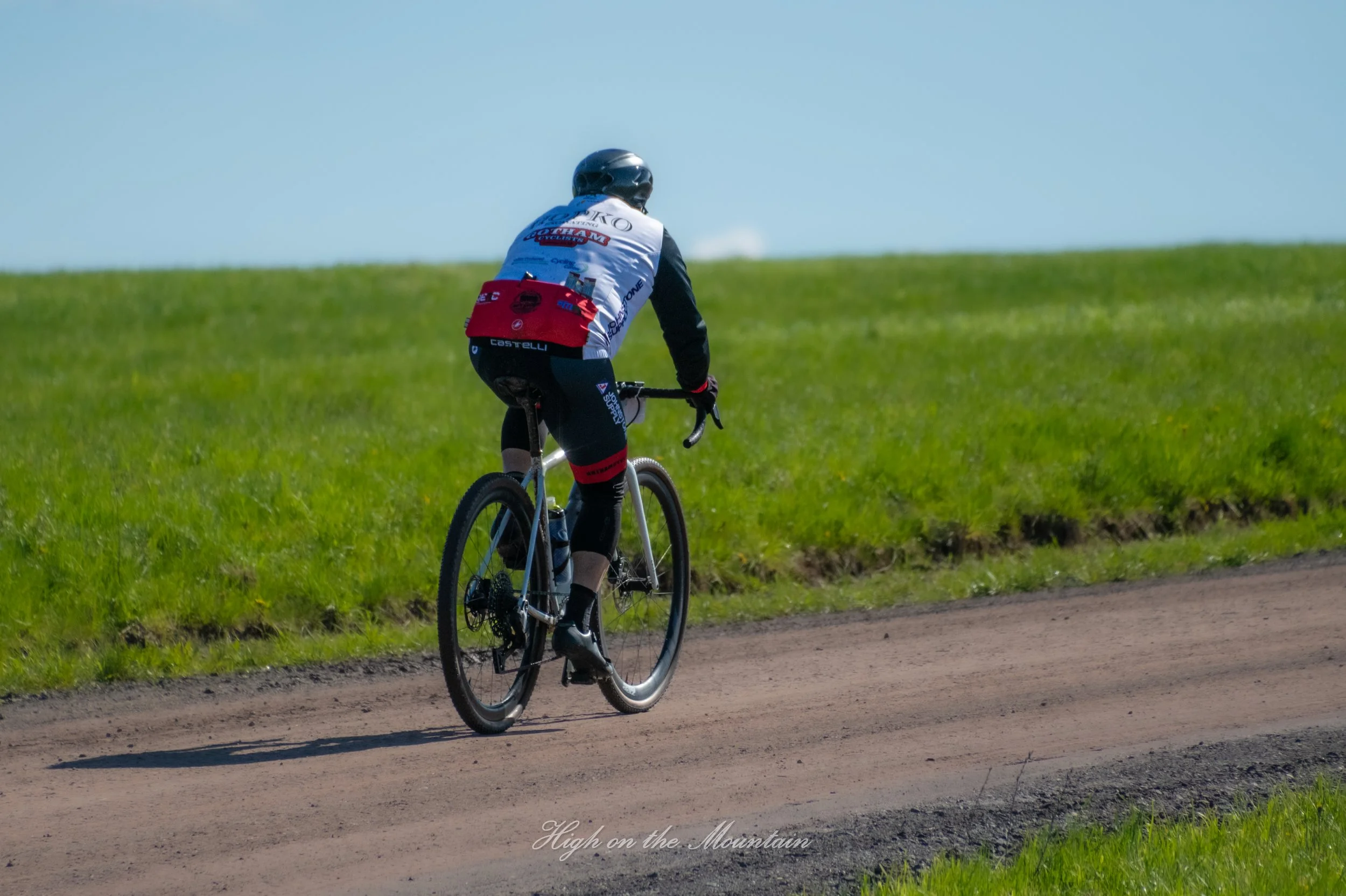 A person rides a bicycle on a dirt path through a green field with blue sky above.