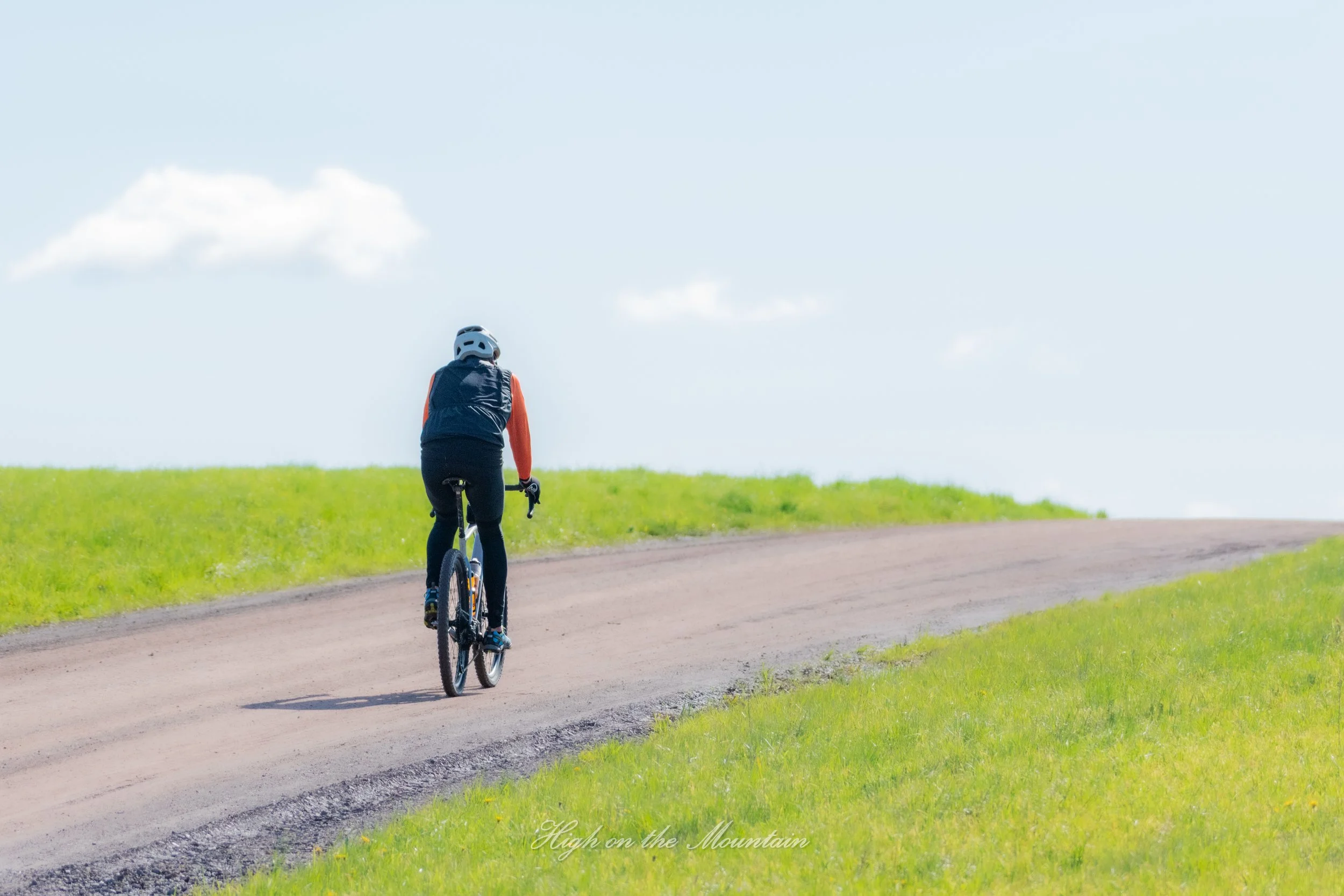 A person riding a mountain bike on a dirt path through a grassy landscape under a partly cloudy sky.