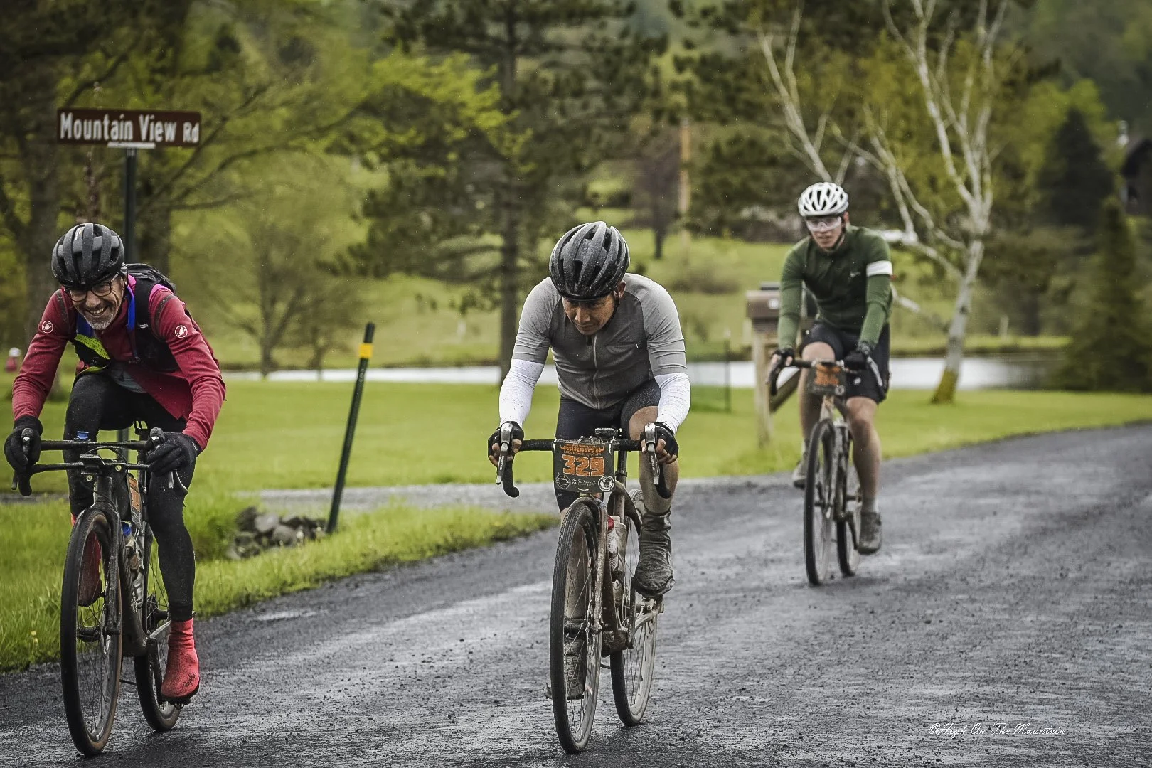 Three cyclists wearing helmets ride their bikes on a gravel path in a wooded area during daytime. They appear to be participating in a race or group ride.