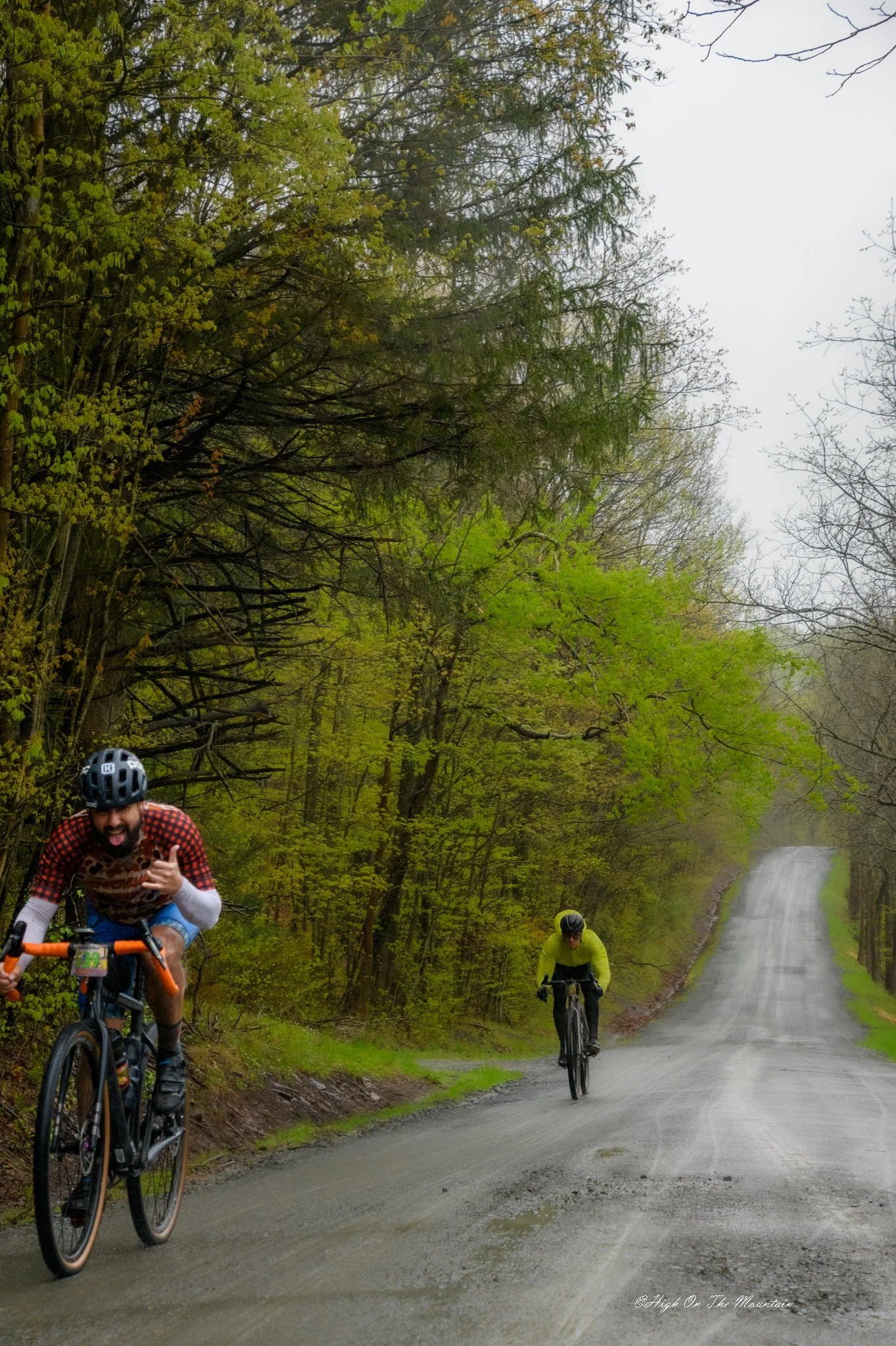 Three cyclists riding on a muddy forest trail, surrounded by green trees and overcast sky, with a waterfall visible in the background.