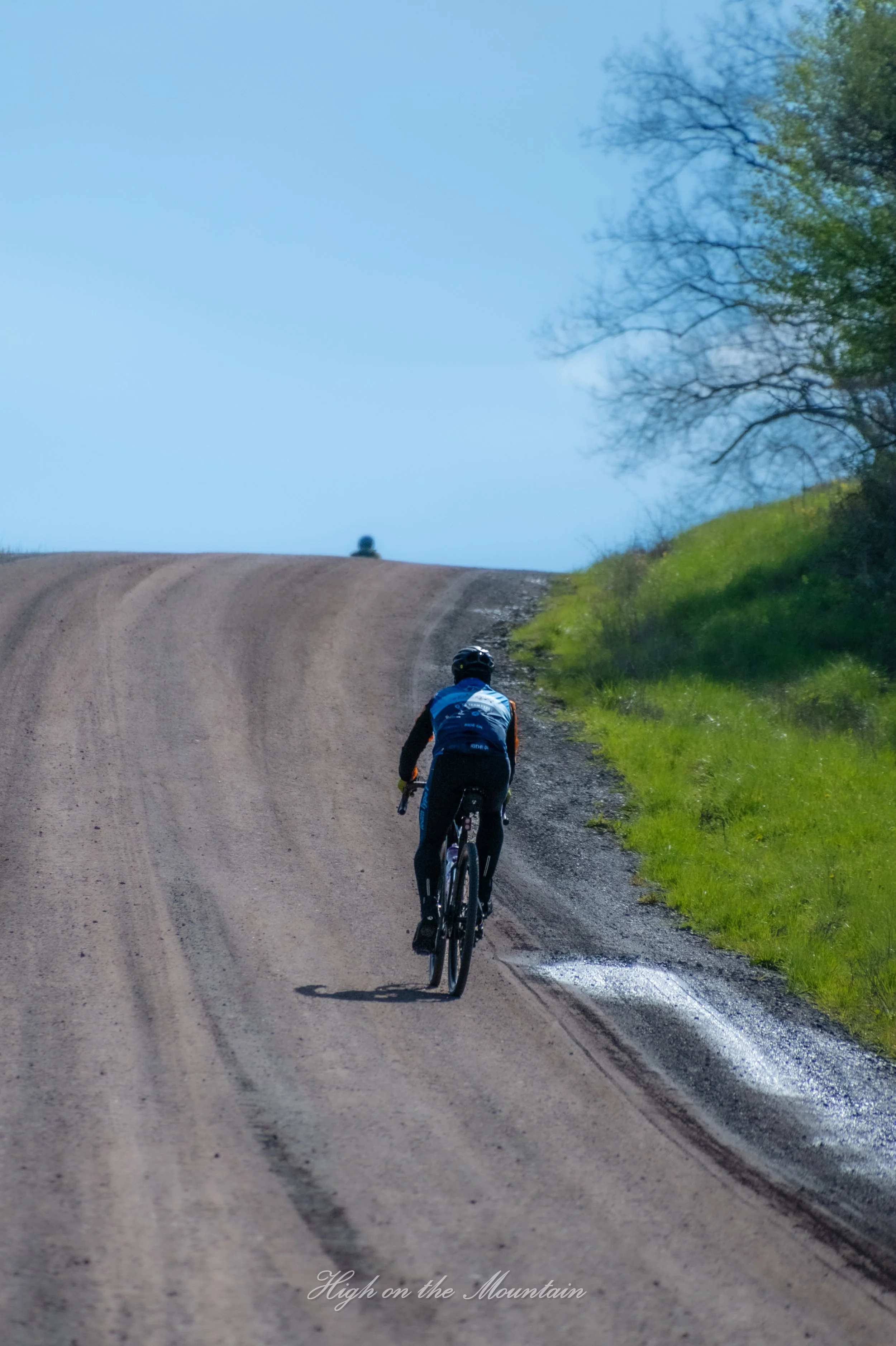 A cyclist riding uphill on a dirt mountain trail surrounded by green grass and trees under a blue sky.