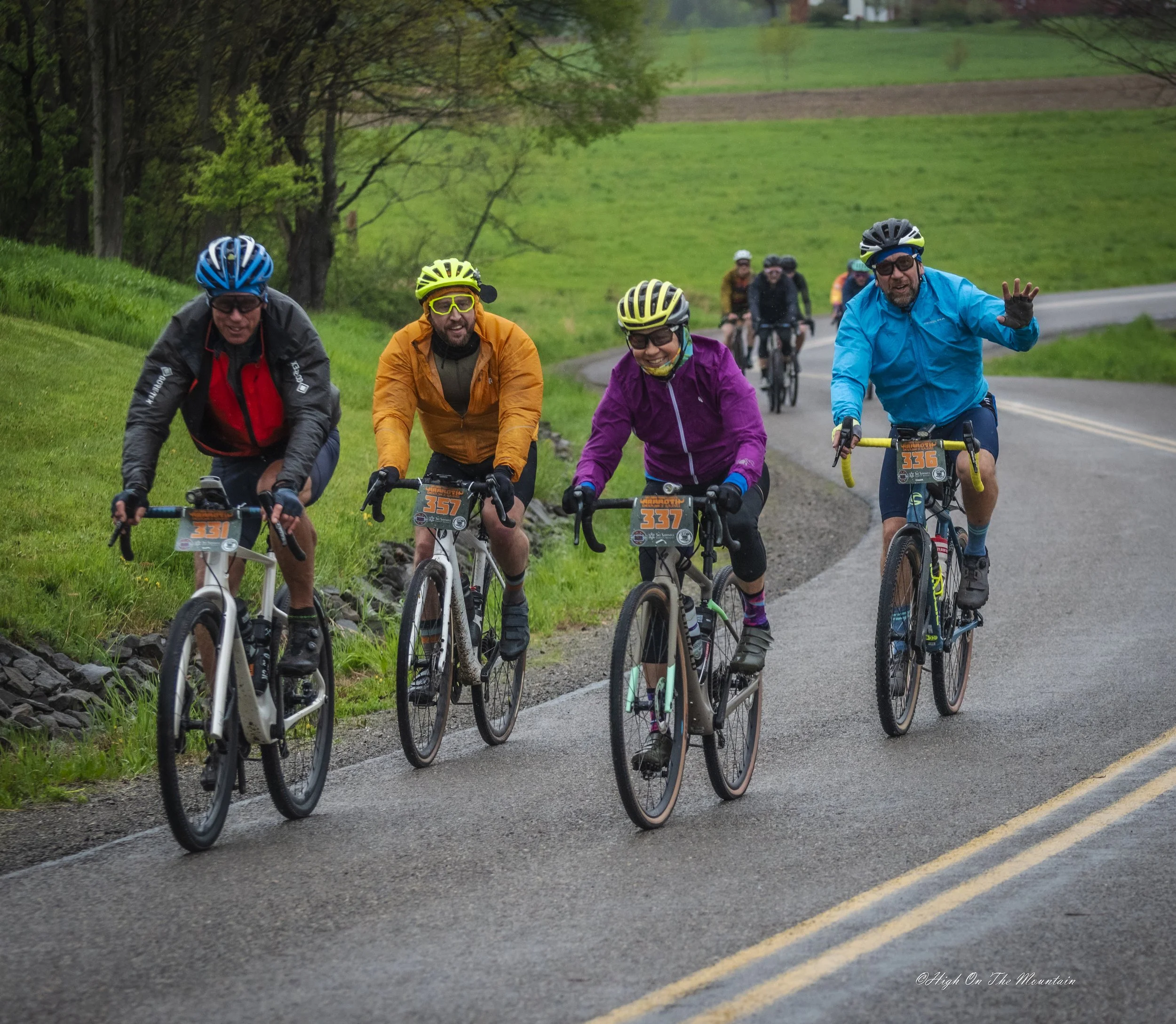A group of seven cyclists riding on a rural road with green fields and trees in the background. They are wearing helmets and colorful jackets, and appear to be enjoying a ride together.