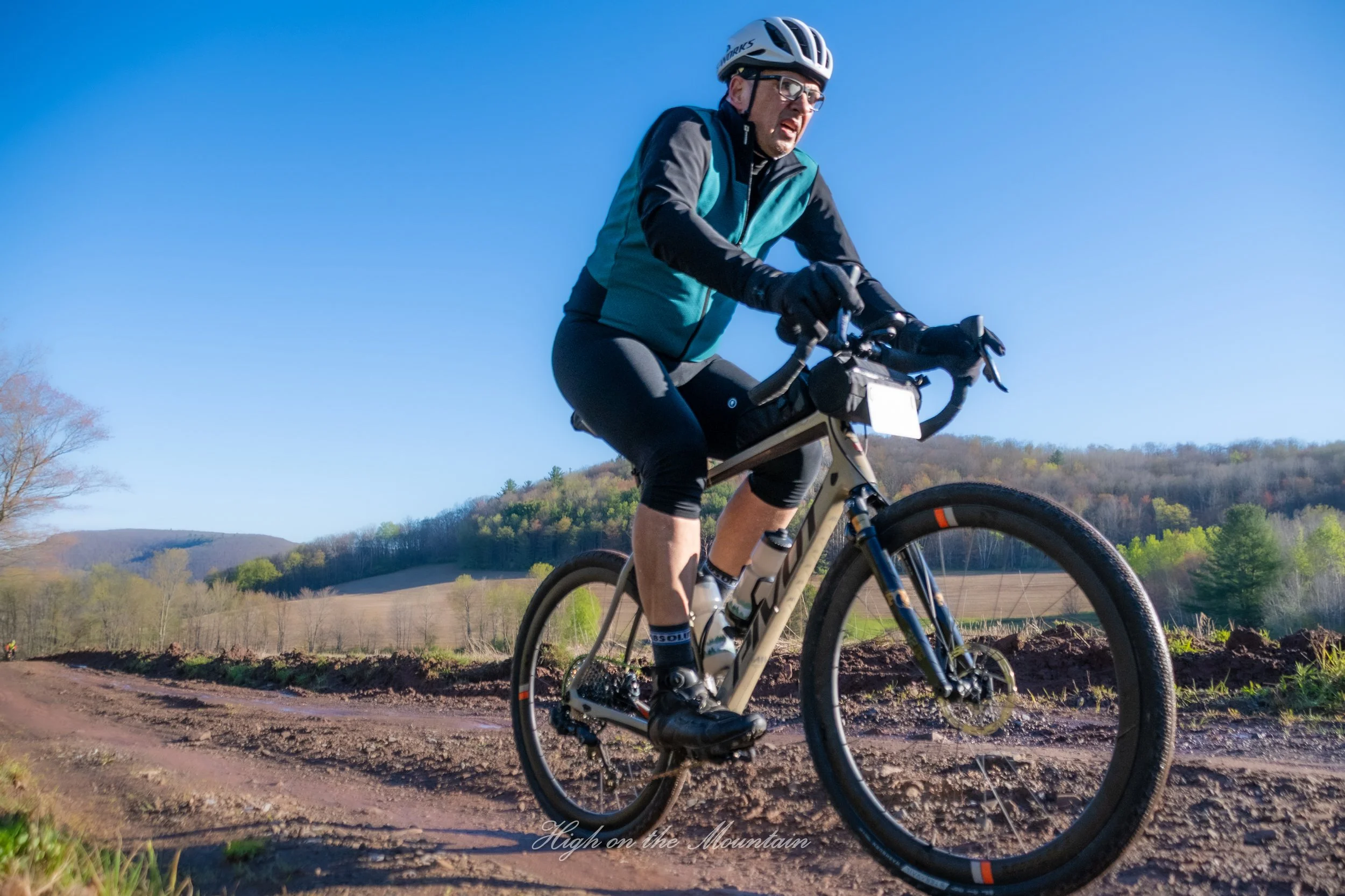 A man riding a mountain bike on a dirt trail in a rural landscape under a clear blue sky. He is wearing a helmet, glasses, and a teal and black cycling outfit. Hills and trees are visible in the background. The text 'High on the Mountain' appears at 