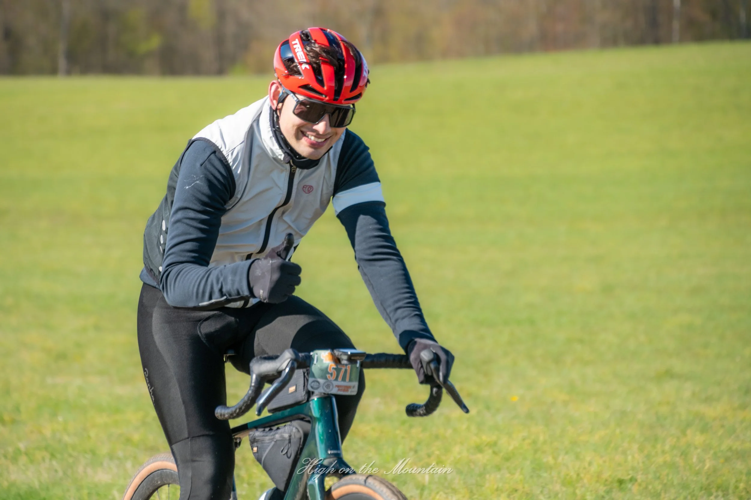 Man cycling outdoors on a sunny day, wearing a red helmet, dark sunglasses, a white and dark jacket, and black pants, smiling and giving a thumbs-up.