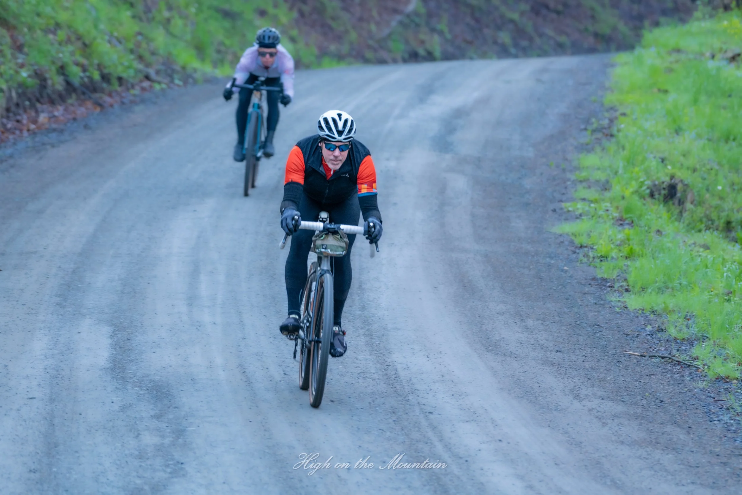 Two cyclists riding on a dirt mountain trail surrounded by greenery.