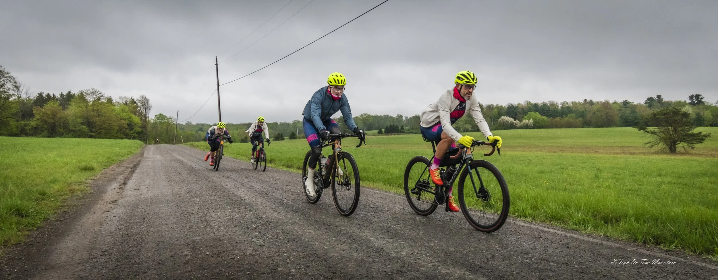 Four people riding mountain bikes on a gravel road through a green field on a cloudy day, wearing helmets and colorful cycling gear.