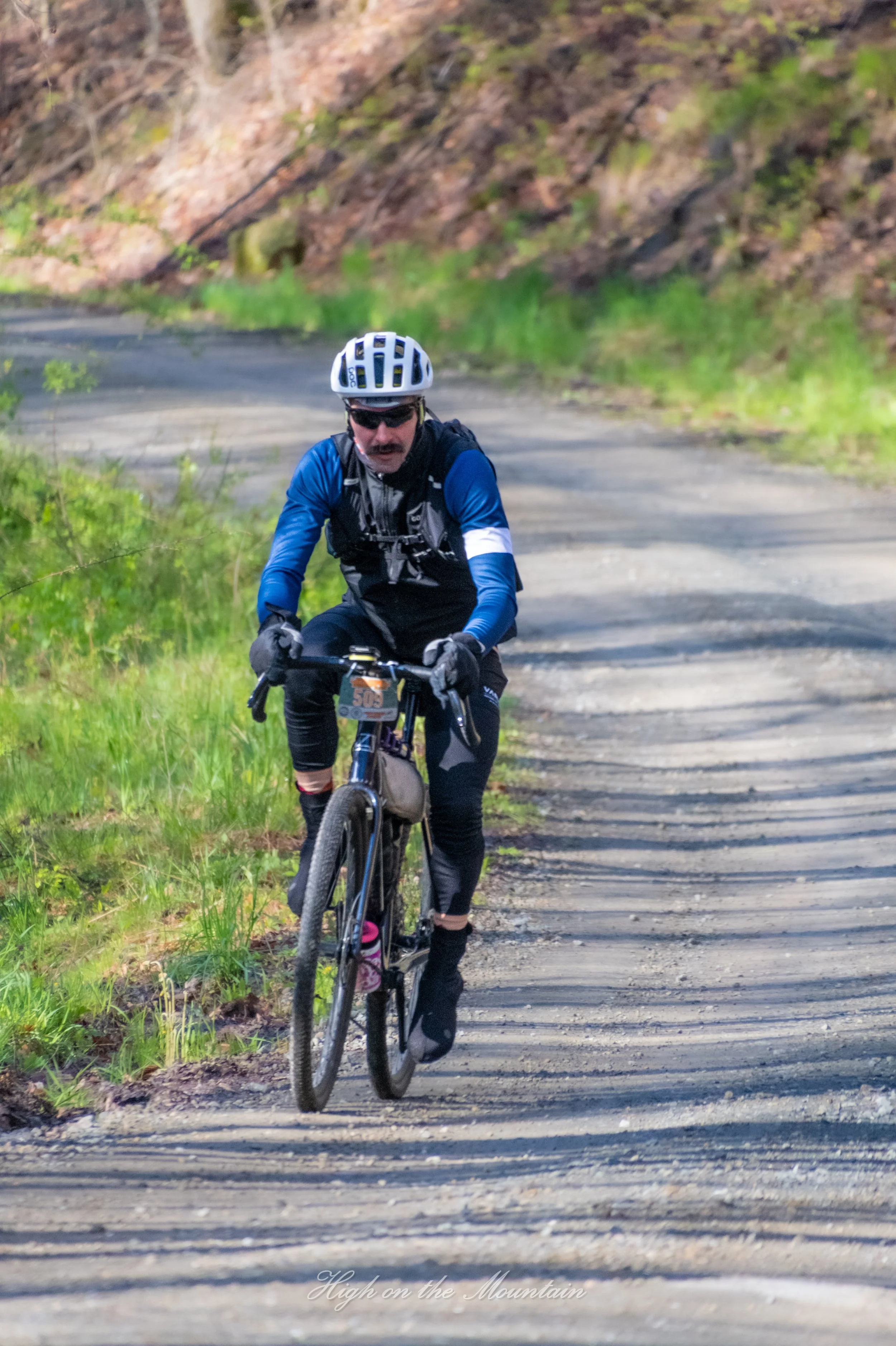 A mountain biker in a blue and black outfit riding on a dirt trail through a lush green forested area. The biker is wearing a white helmet, sunglasses, and a hydration pack. There is a text overlay that reads 'High on the Mountain'.