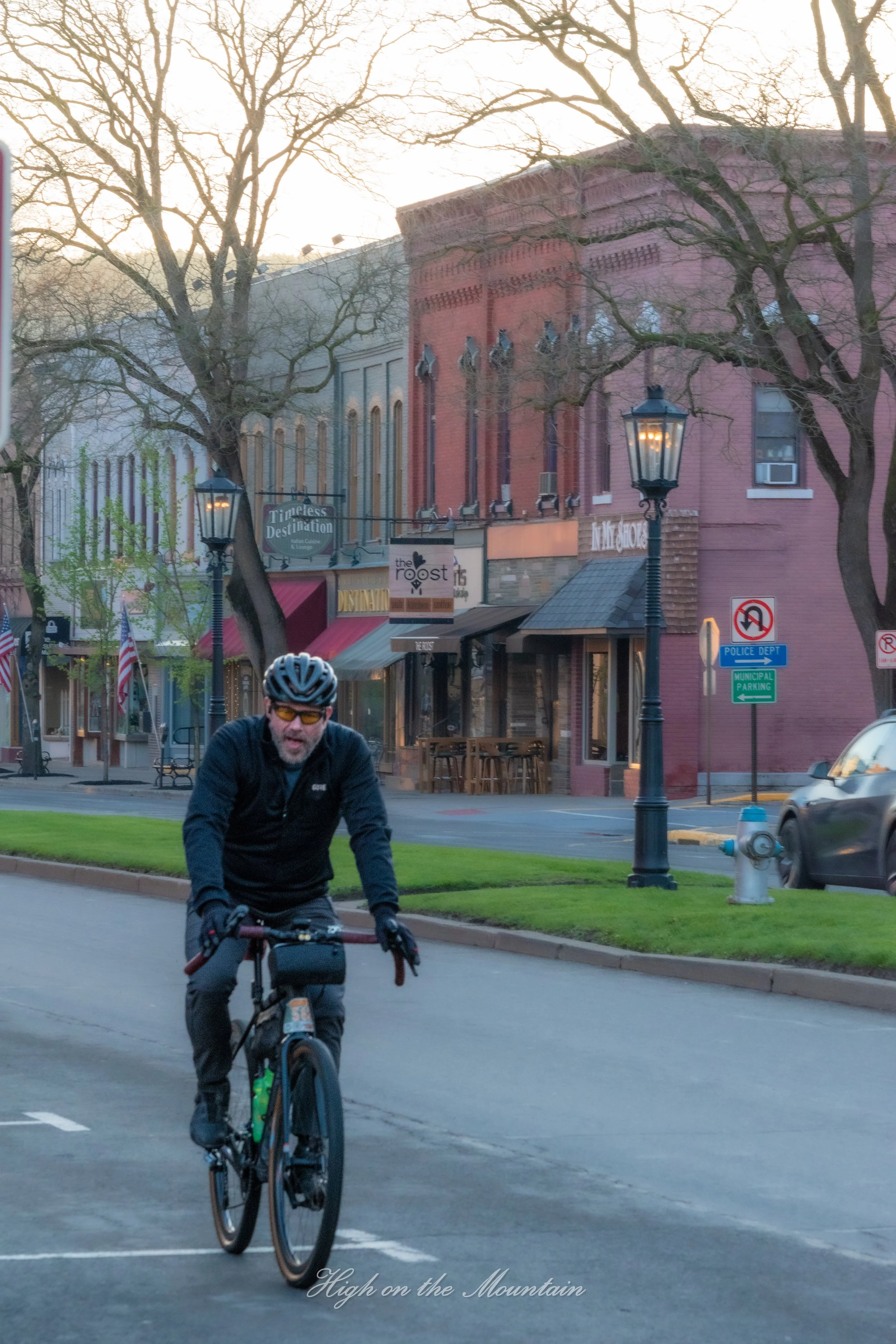 A man riding a bicycle in a parking lot near a grassy area, with buildings, trees, street lamps, and signs including a police department sign in the background.