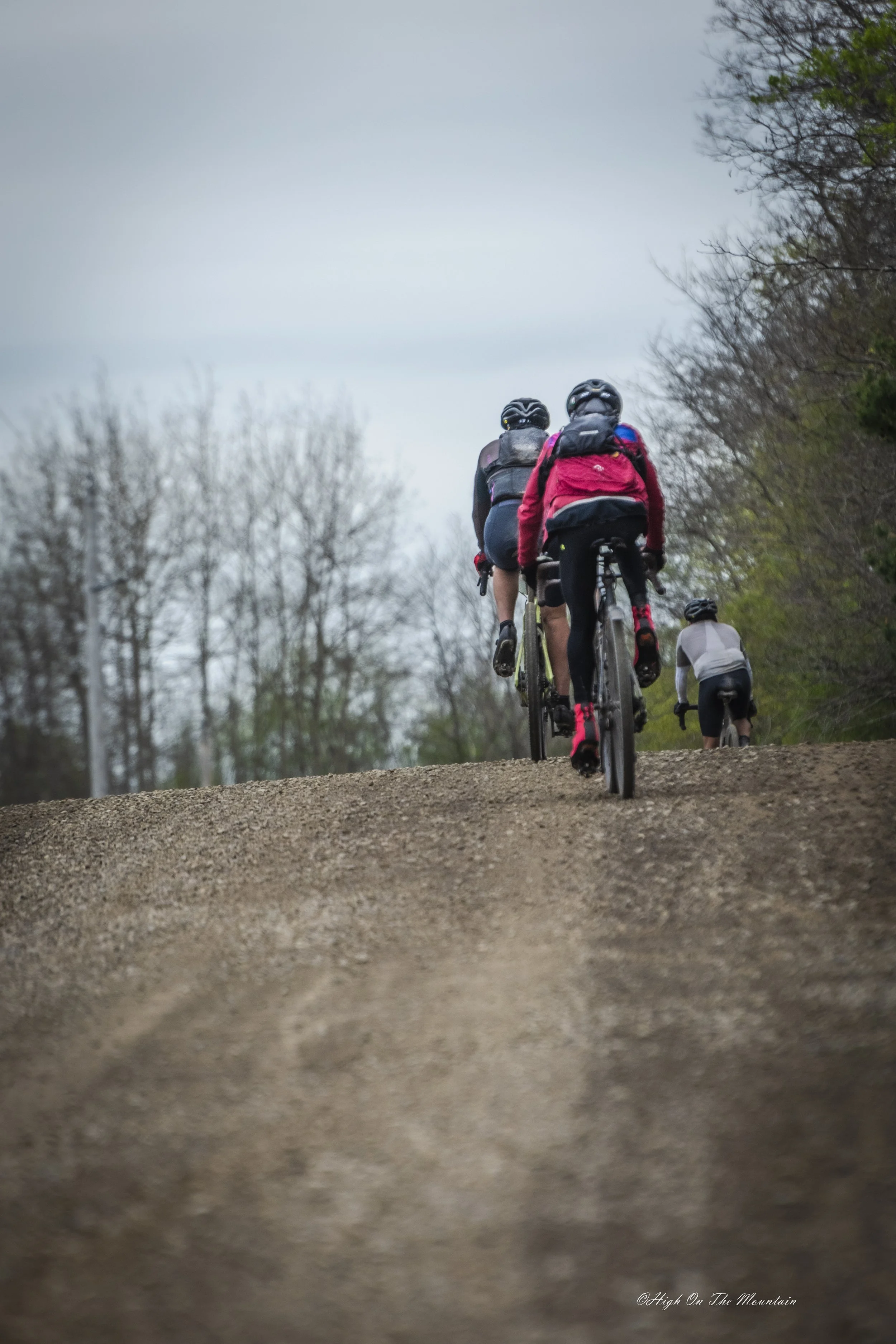 Four cyclists wearing helmets and backpacks riding uphill on a gravel trail surrounded by trees.