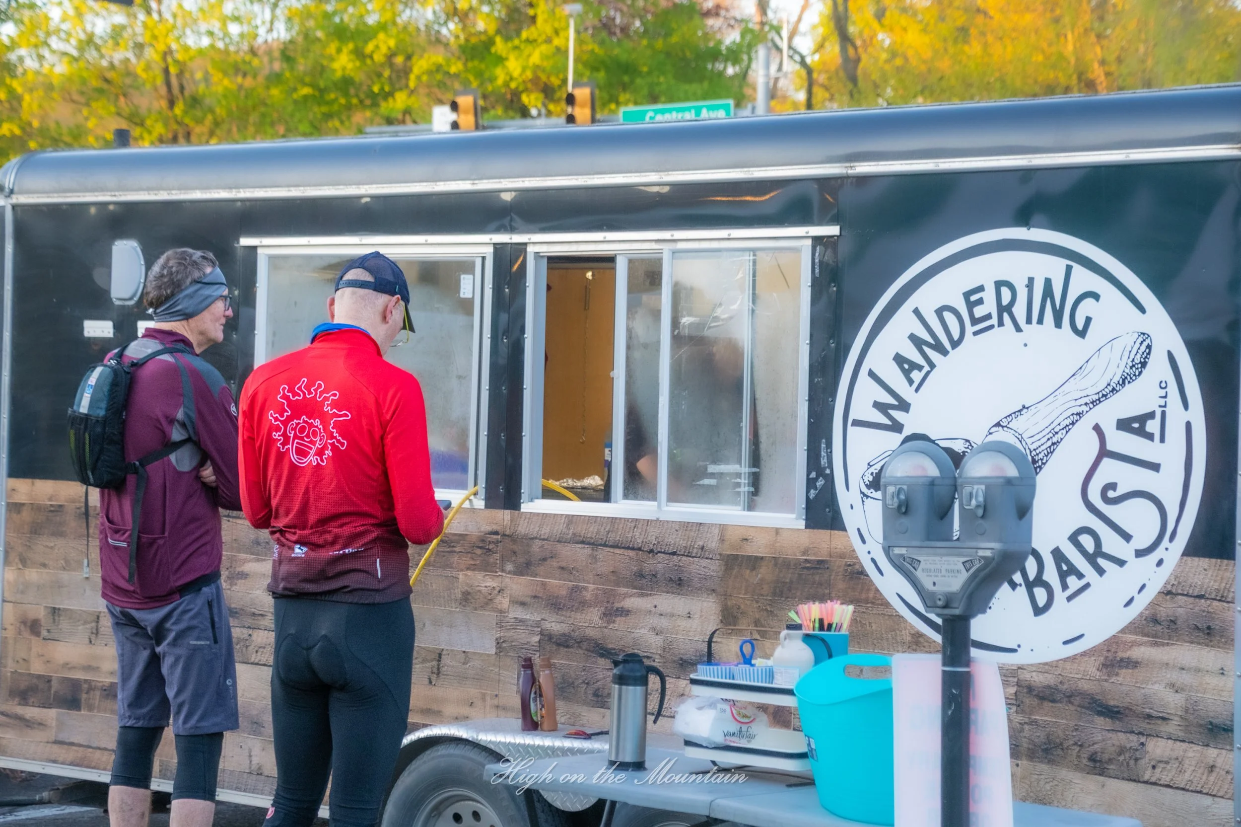 Two people stand in front of a black food truck with a logo featuring a fishing pole. The food truck is parked outdoors on a sunny day, with trees showing autumn foliage in the background. The people are looking inside the food truck window, possibly