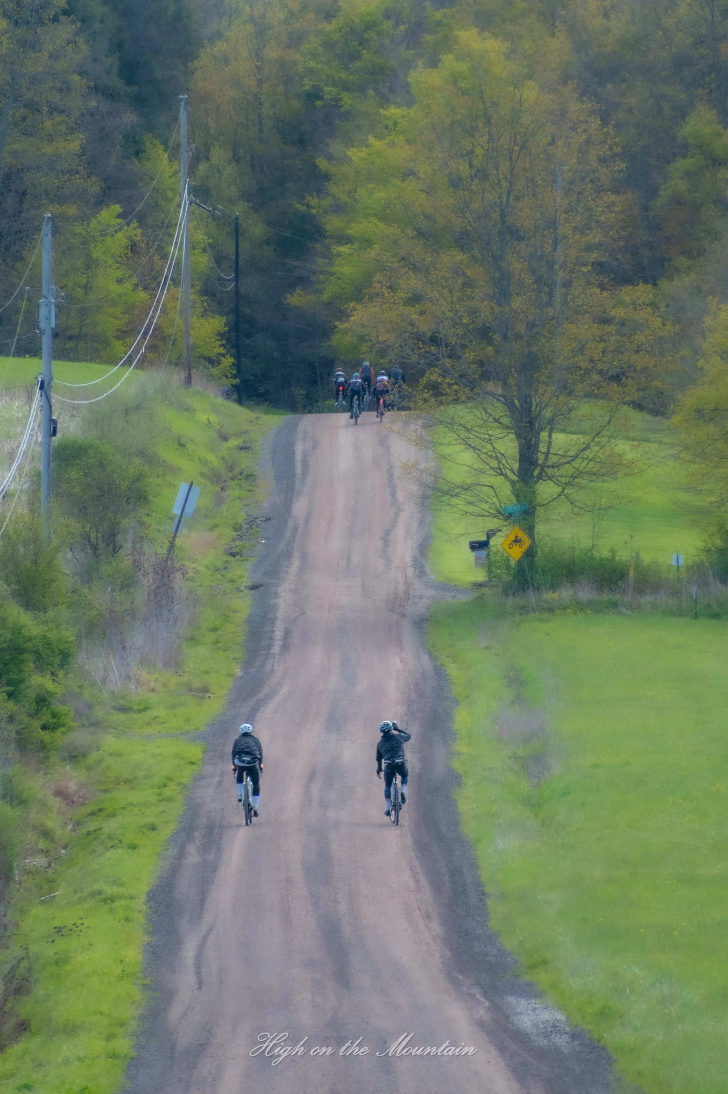 A group of cyclists riding along a dirt trail in a lush, green, hilly area with trees on either side and utility poles on the left. The sky is overcast, and there is a small yellow sign with a bike symbol on the right.