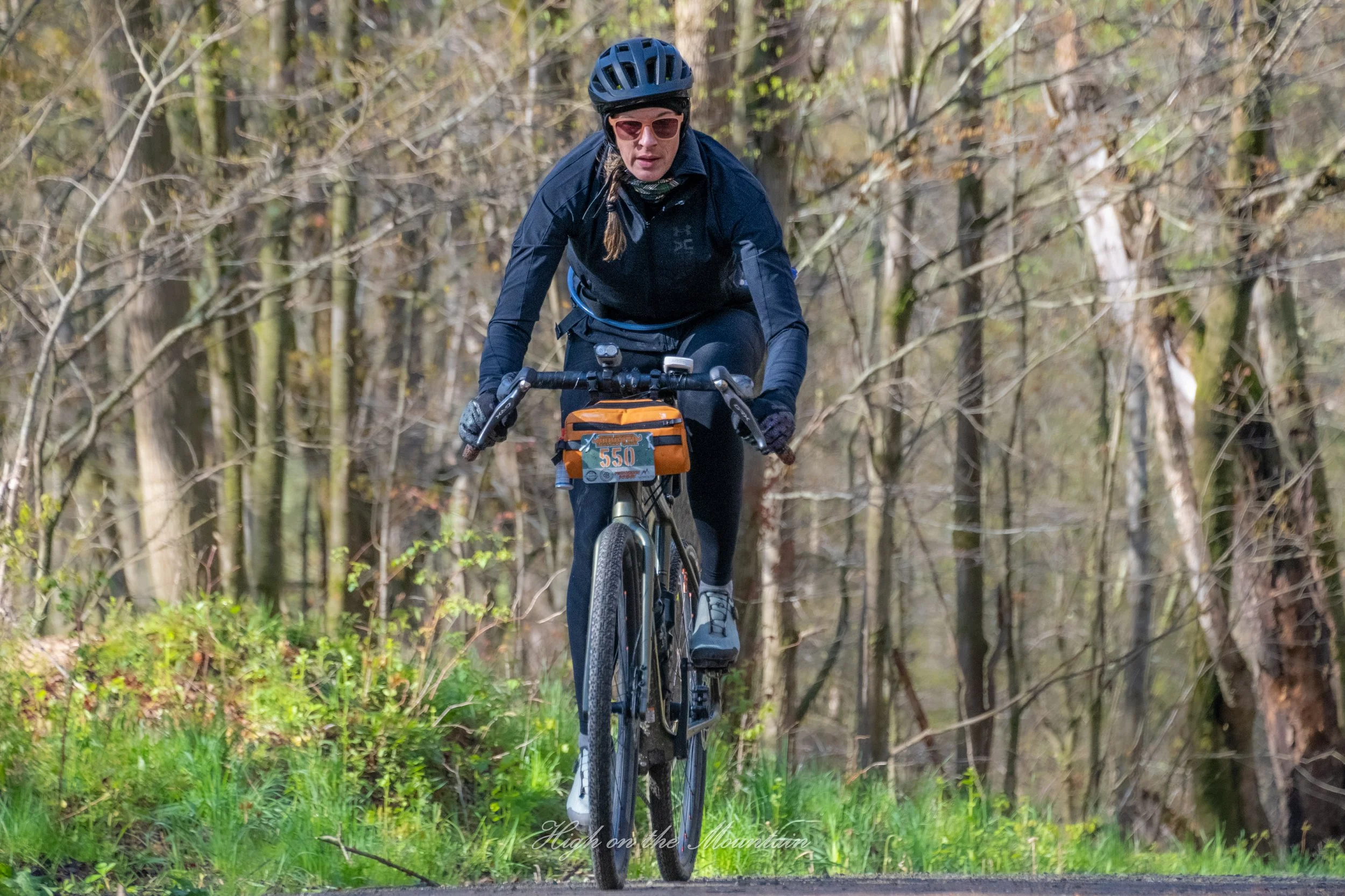 A woman wearing a helmet and sunglasses riding a mountain bike through a forested trail with trees and fallen leaves.
