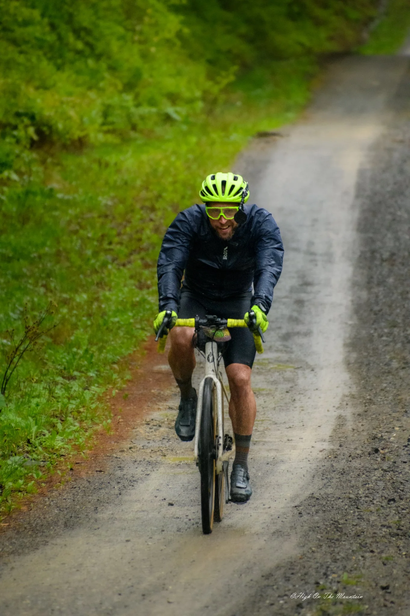 A man riding a mountain bike on a dirt trail through a forested area with green foliage, wearing a bright yellow helmet, yellow glasses, a black jacket, shorts, and cycling shoes.