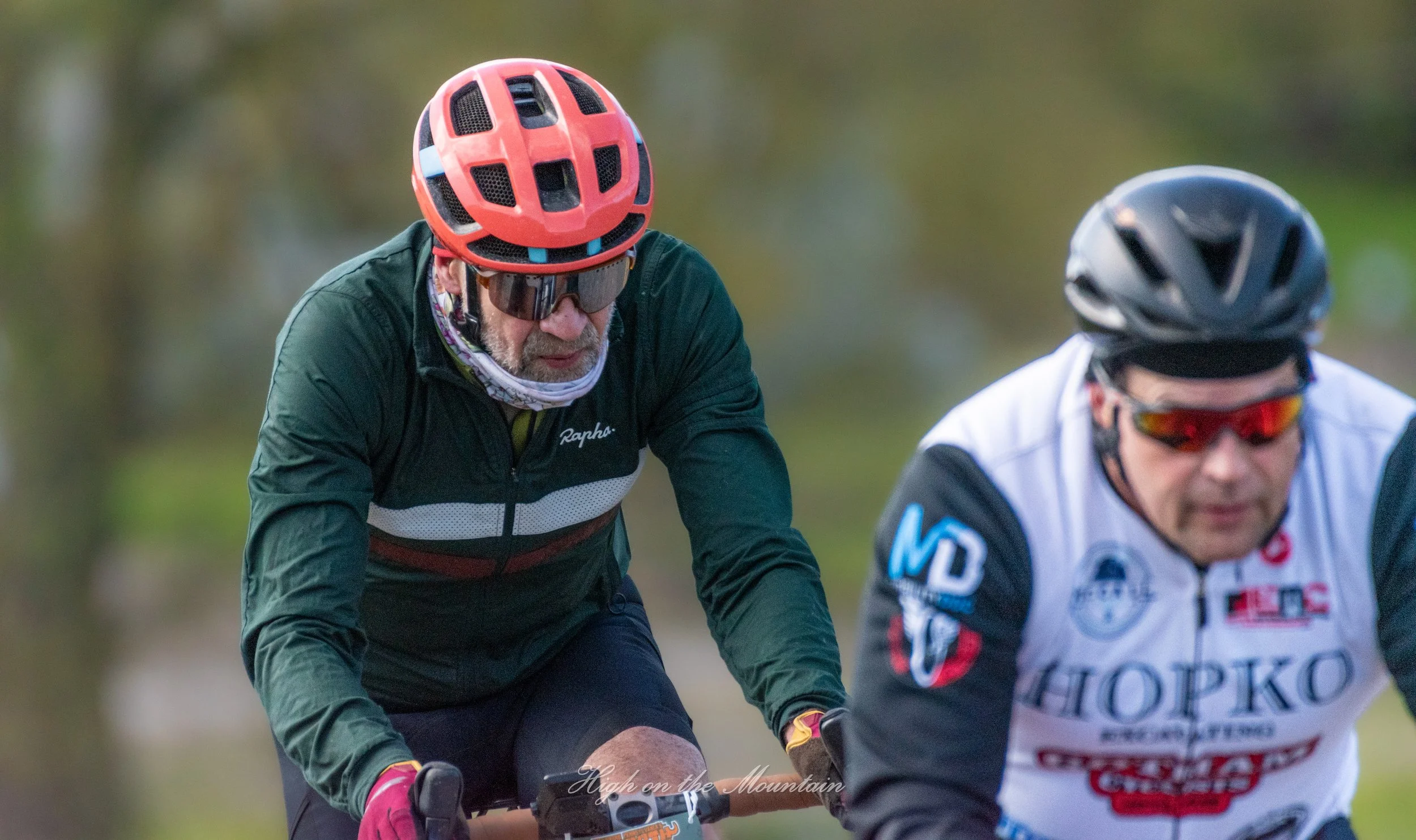 Two cyclists riding outdoors, one wearing a red helmet and sunglasses, and the other wearing a black helmet and sunglasses, with blurred green background.