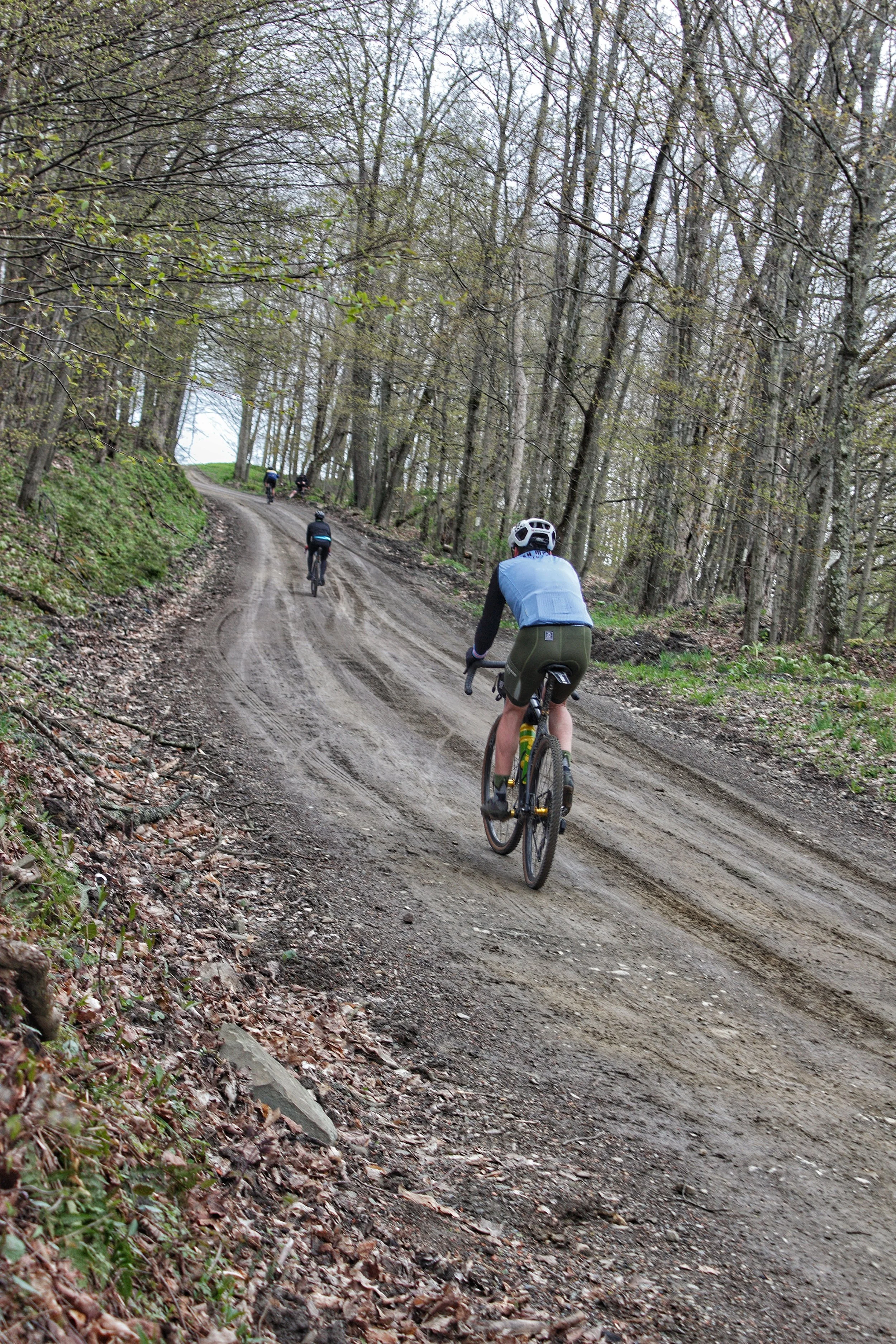 Group of cyclists riding on a dirt trail through a forest with leafless trees.