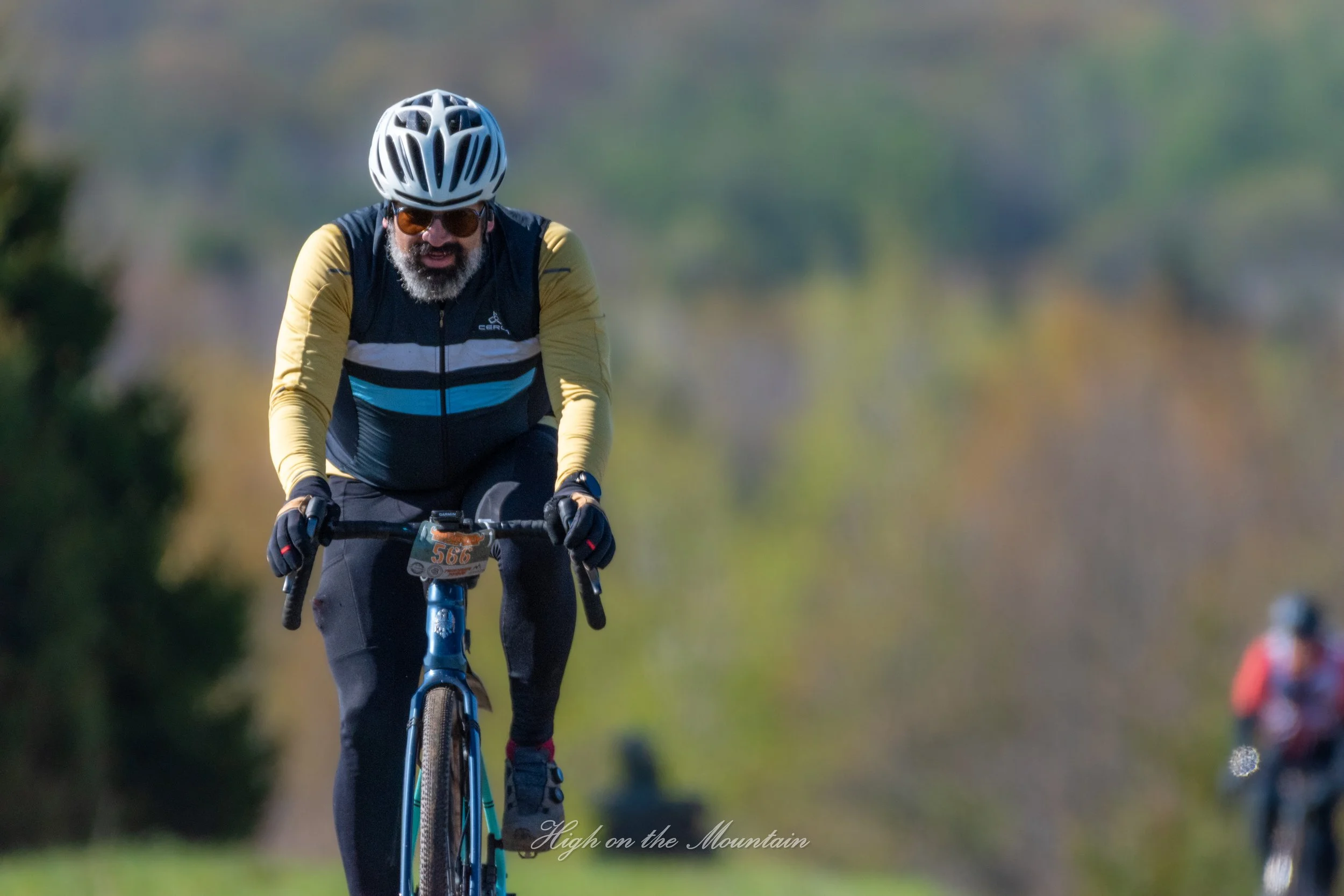A man with a beard and sunglasses riding a mountain bike outdoors during daytime, wearing a helmet, yellow and black cycling jersey, and black pants, with a blurred natural background.