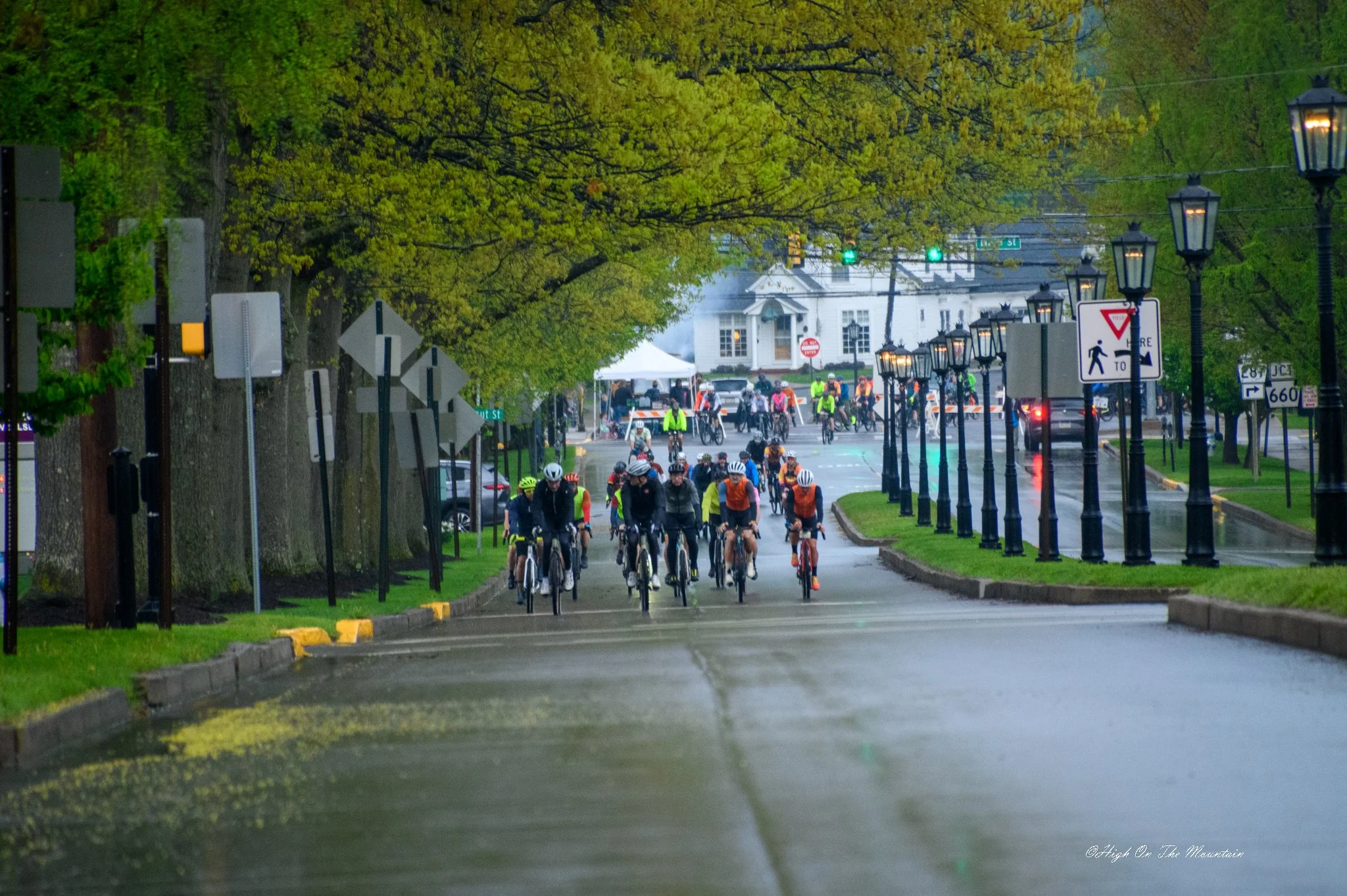 Bicyclists riding along a street lined with large green trees and vintage black lampposts, with a white house and traffic lights in the background on a rainy day.