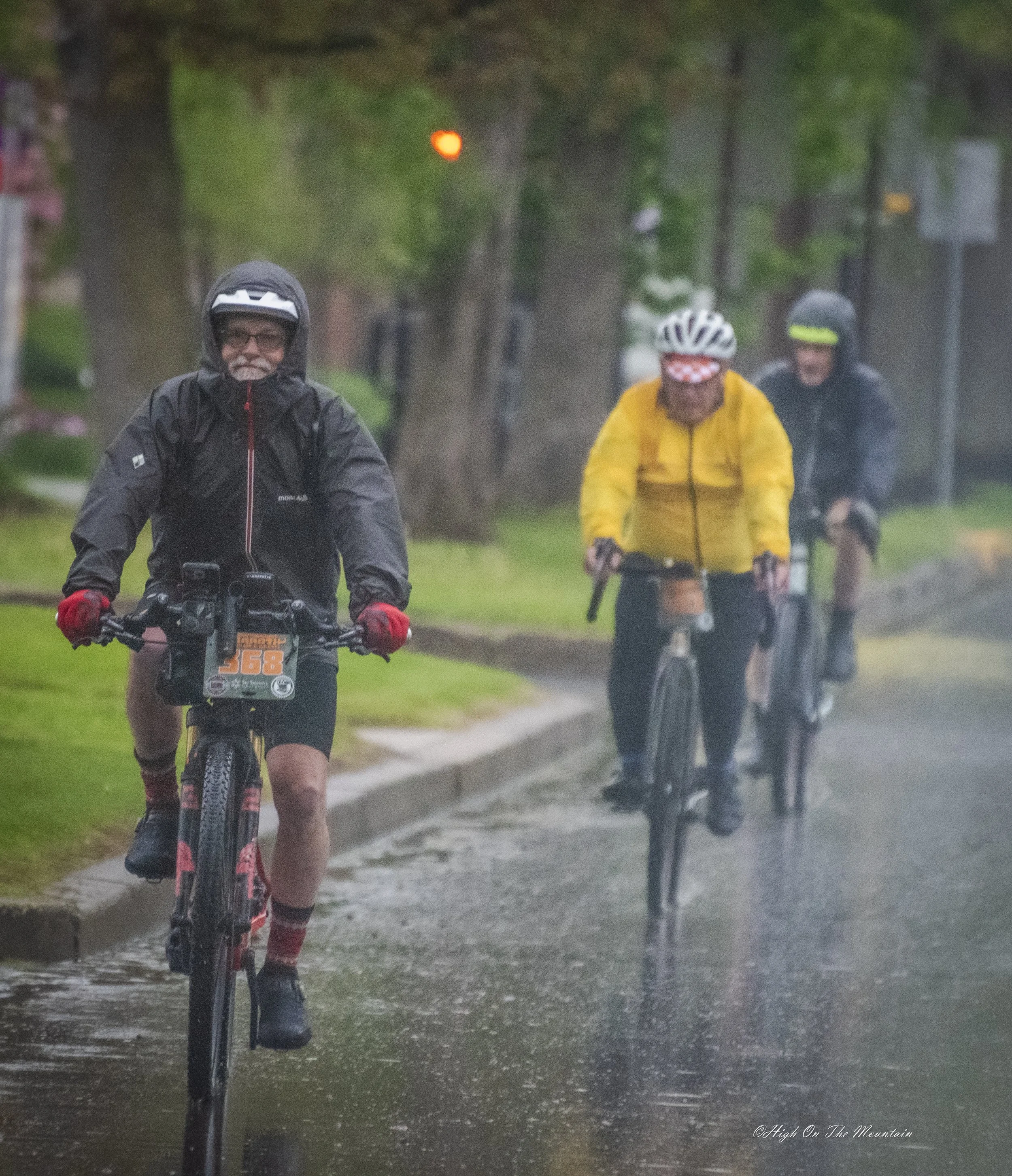 Three cyclists riding in the rain on a tree-lined street, wearing rain jackets and helmets, with the water on the ground reflecting their figures and creating a blurry background.