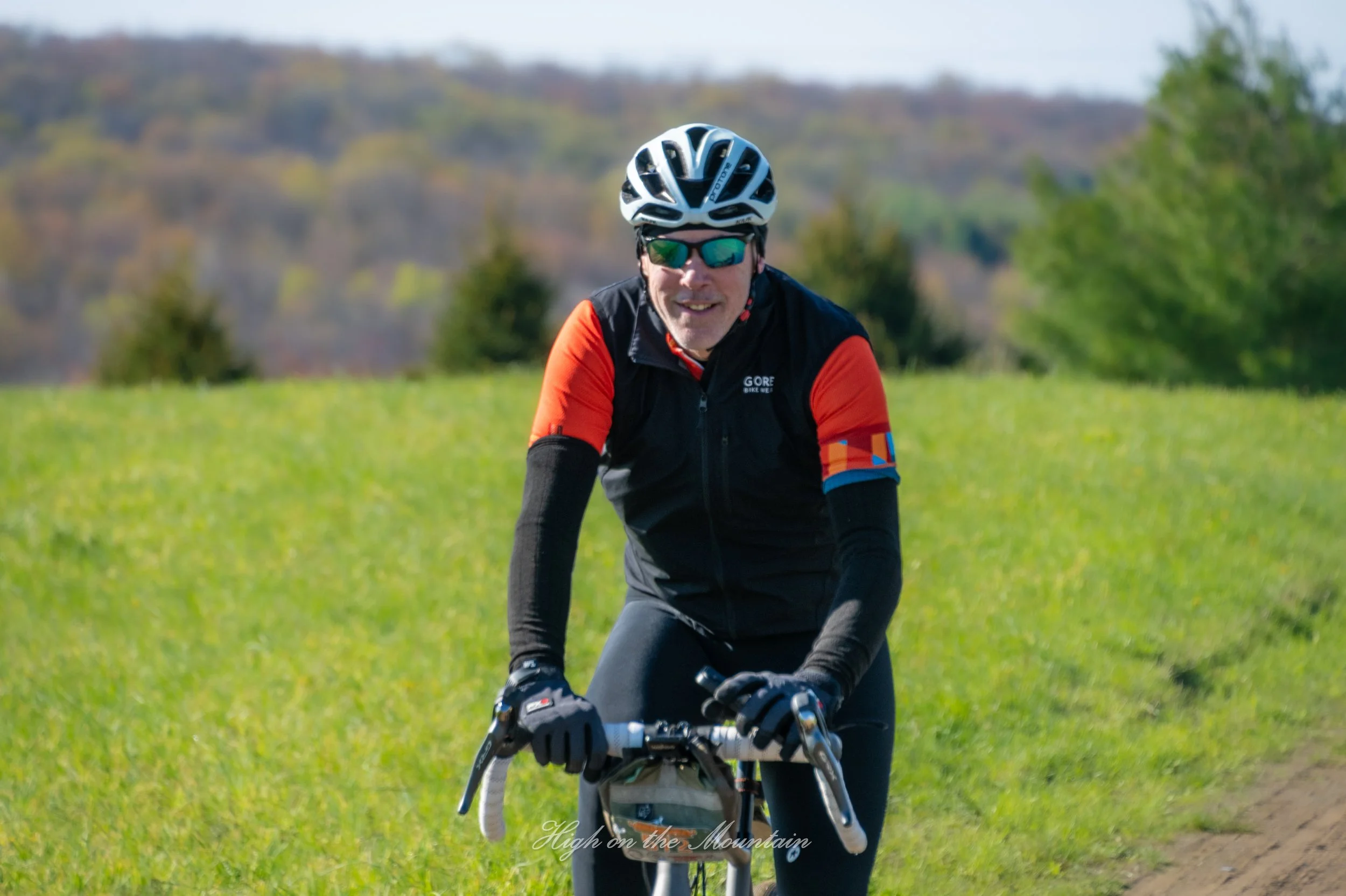 A man riding a mountain bike outdoors on a grassy trail, wearing a helmet, sunglasses, and a black and red cycling jersey.